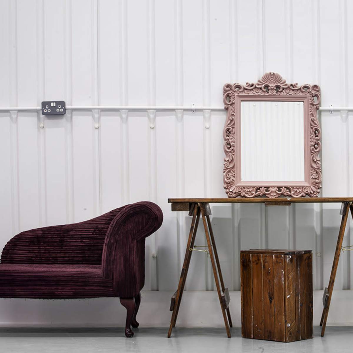 A pink-framed mirror hangs above a wooden table supported by trestles, flanked by a plush couch and a wooden chair against a white wall.