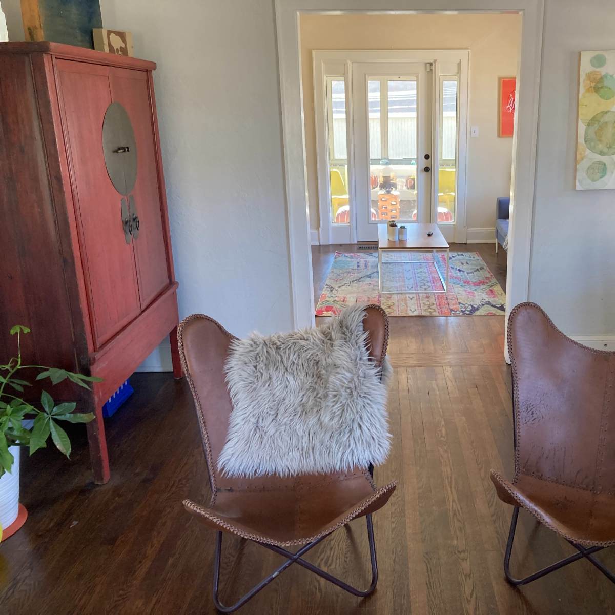 Two leather butterfly chairs with a throw pillow face a hallway leading to a brightly lit room with a colorful rug and doorway.
