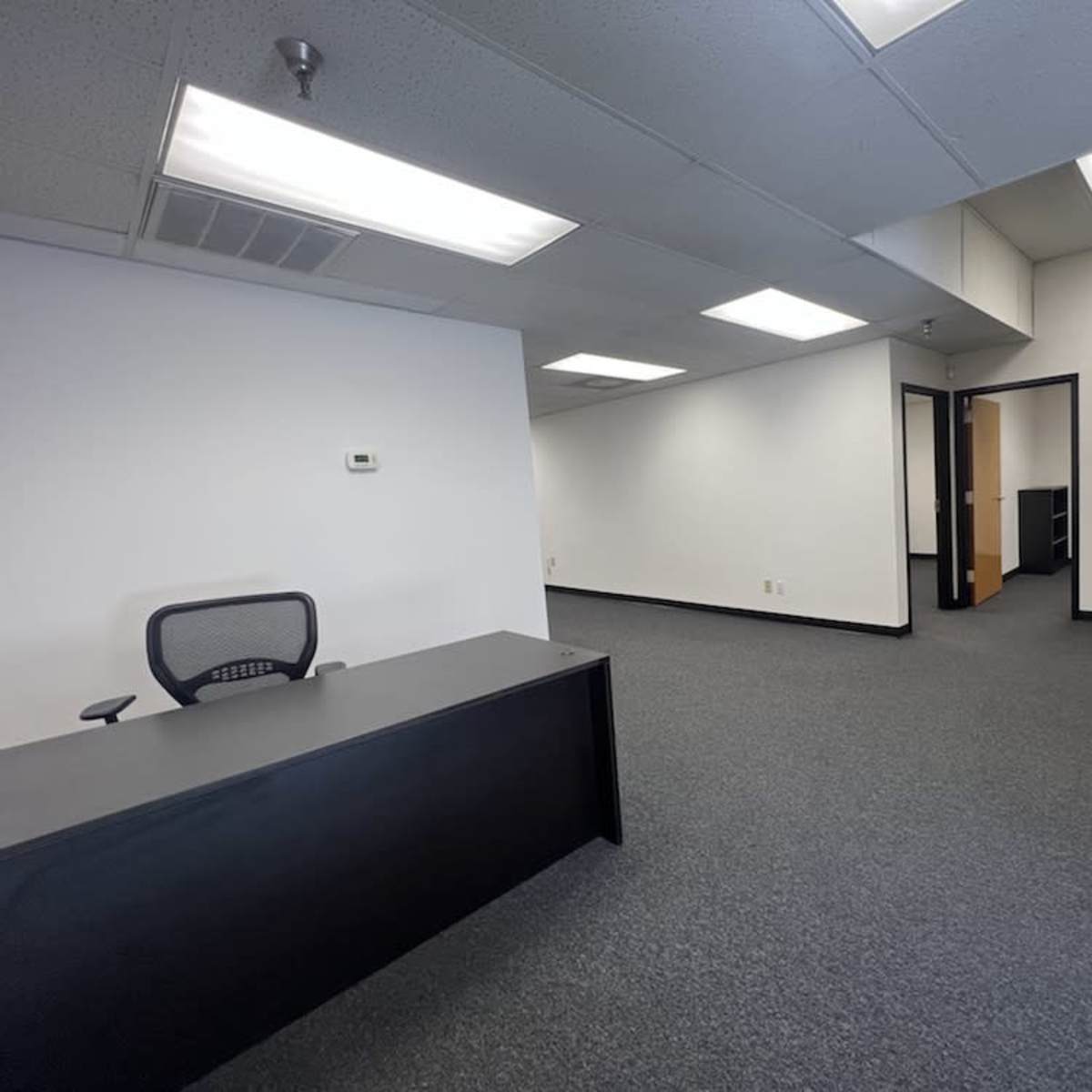 An empty office space with a black reception desk and a waiting area featuring two chairs, all situated on a carpeted floor under simple fluorescent lighting.