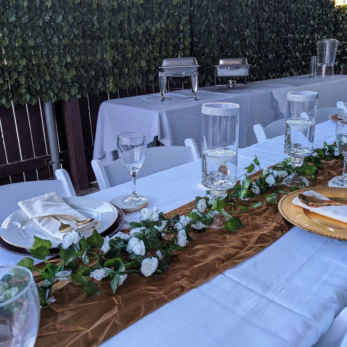 A decorated dining table features white tablecloths, elegant glassware, and a gold runner adorned with greenery and flowers.