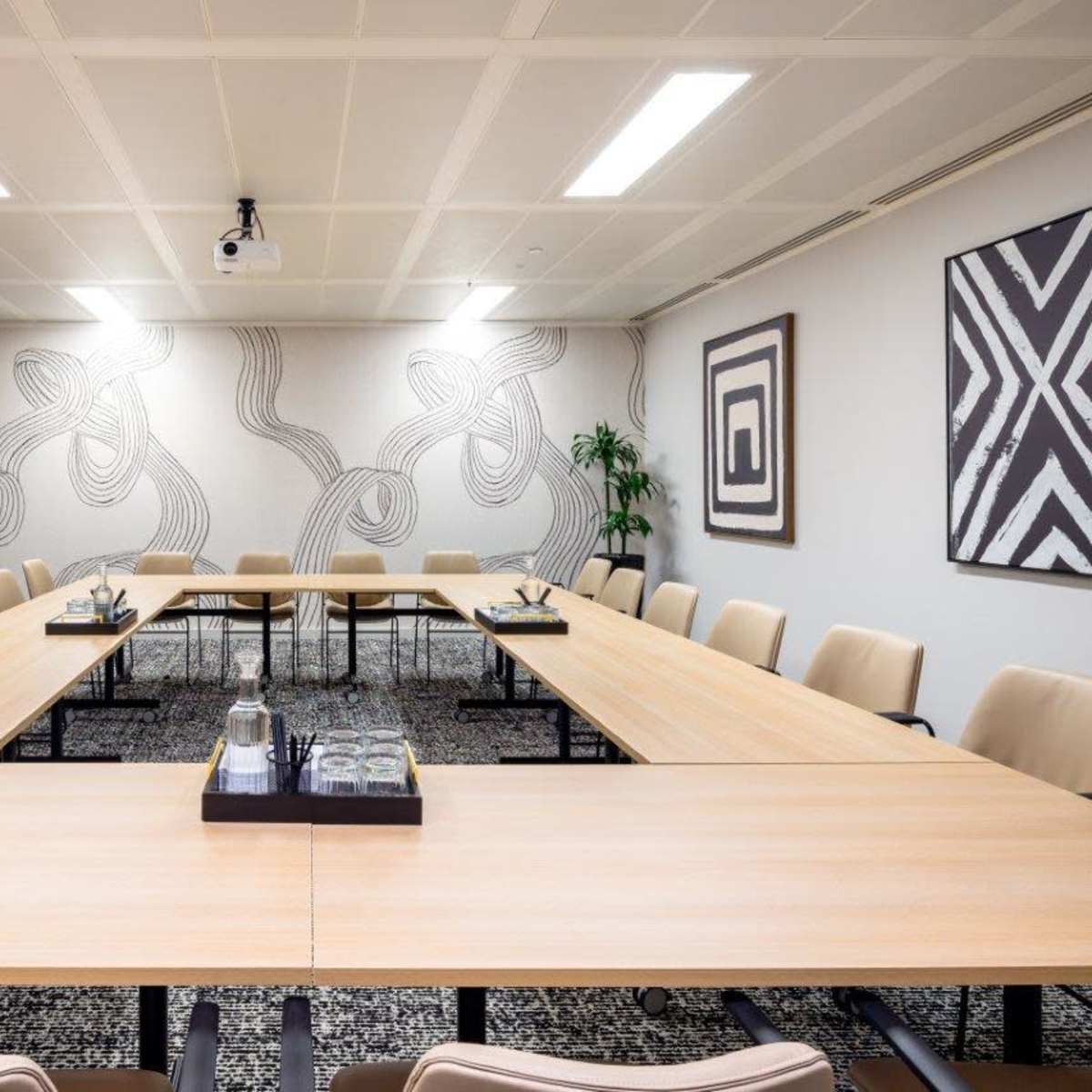 A conference room features a large rectangular table surrounded by beige chairs, with decorative wall art and a projector overhead.