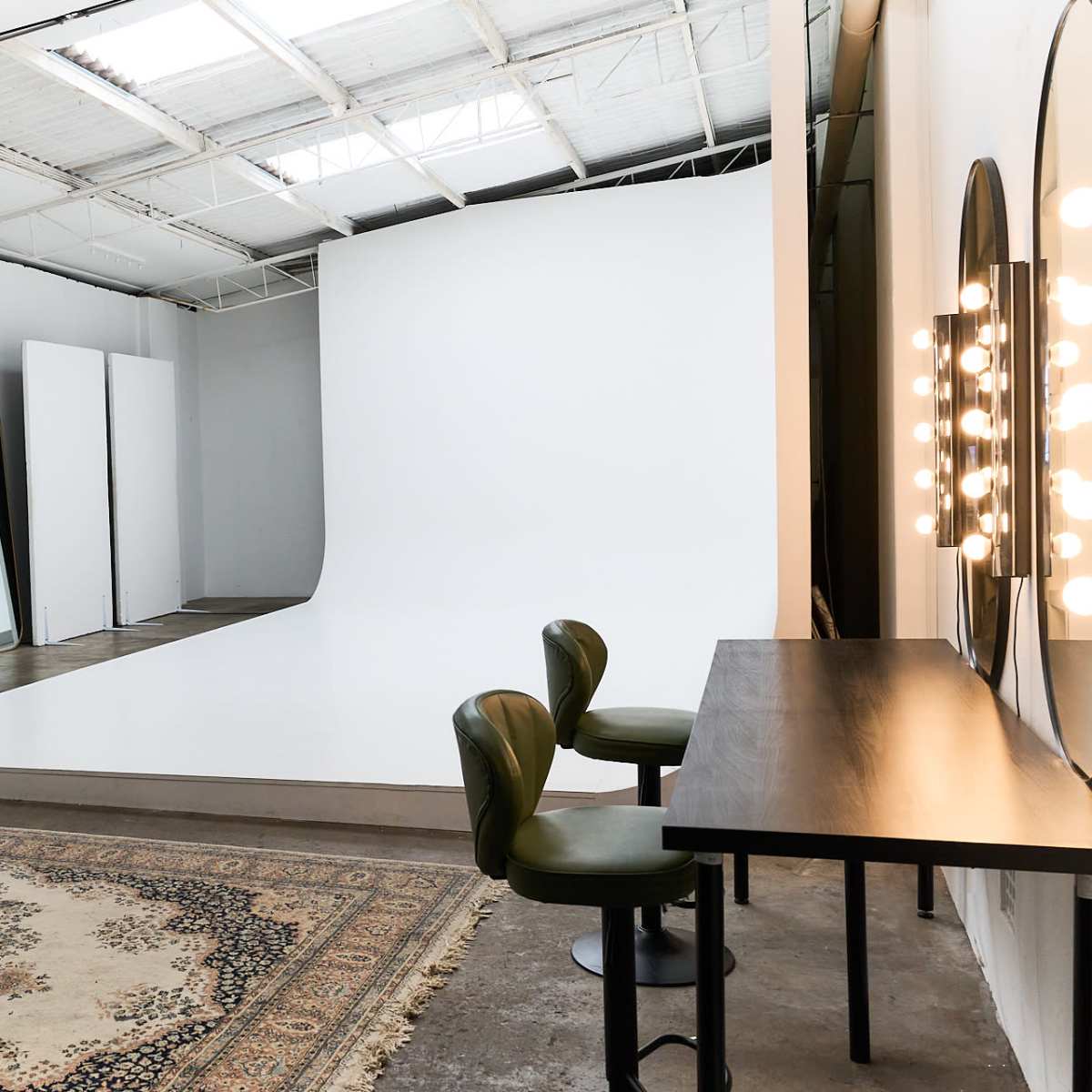 A spacious photography studio featuring a seamless white backdrop, a wooden table with two green chairs, and mirrors with light bulbs, alongside several reflective panels.
