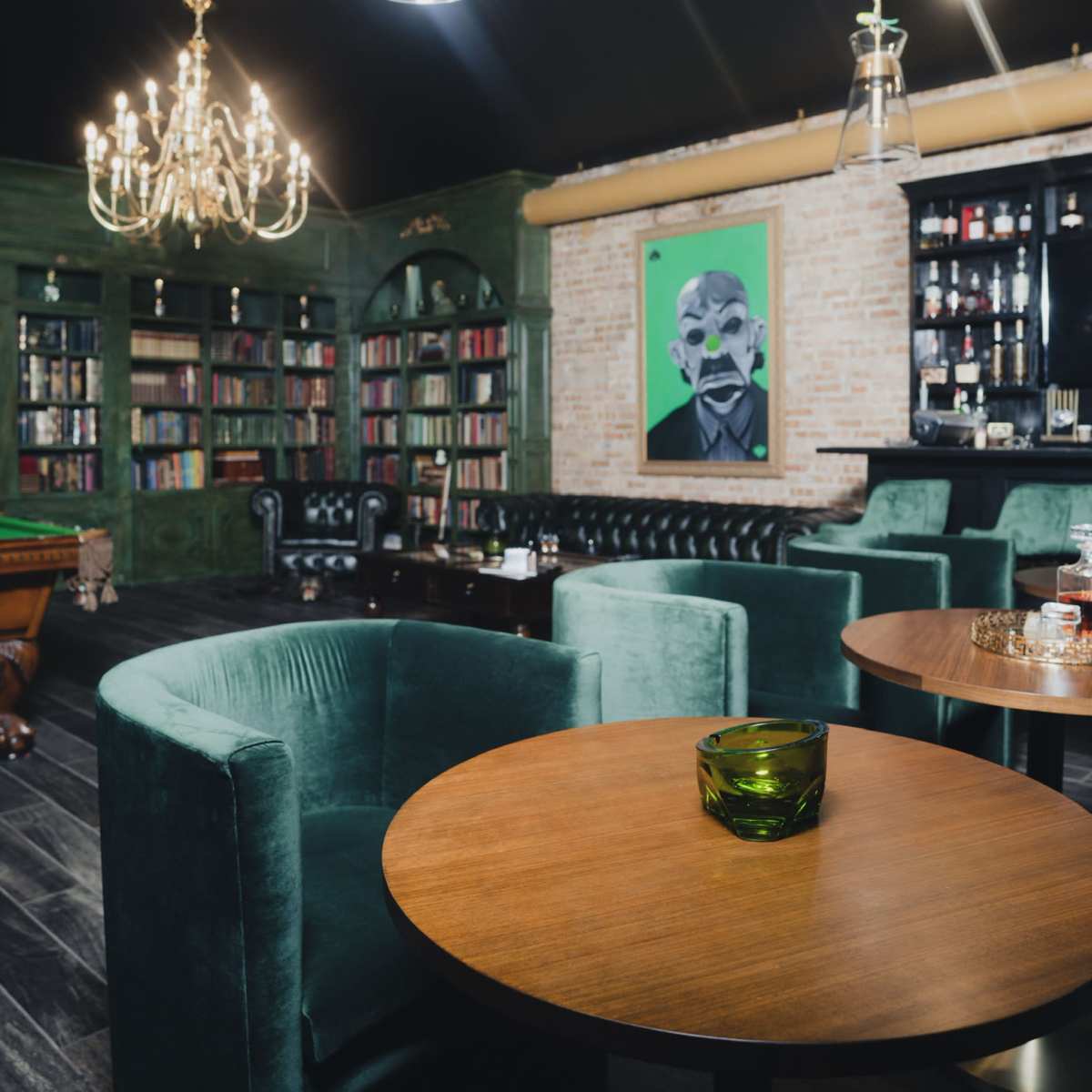 A stylish interior of a bar or lounge with green upholstered chairs, wooden tables, a chandelier, bookshelves, and a vibrant artwork on the wall.