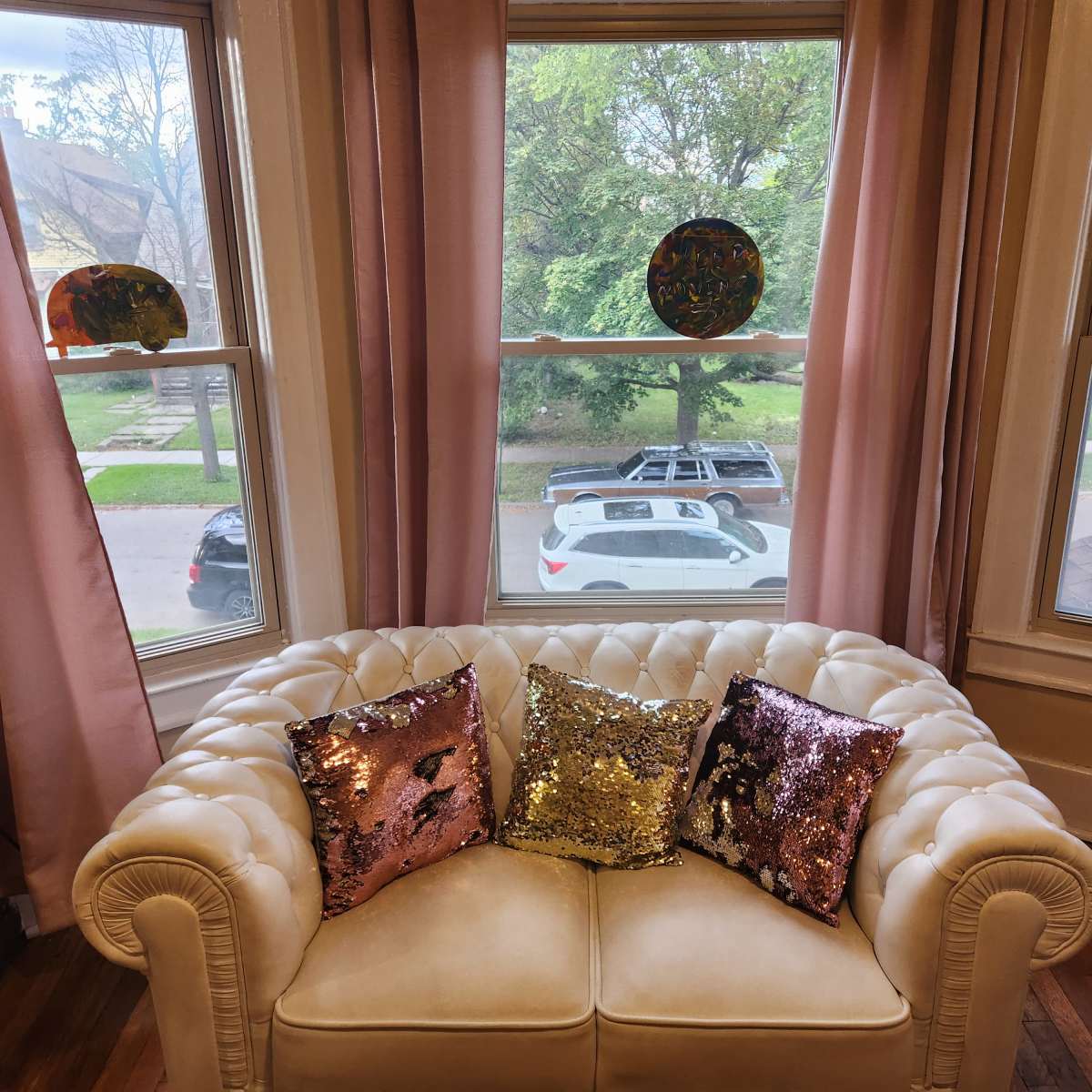 A cream-colored couch with sequined pillows is positioned in front of large windows, showcasing a view of a tree-lined street and parked vehicles outside.