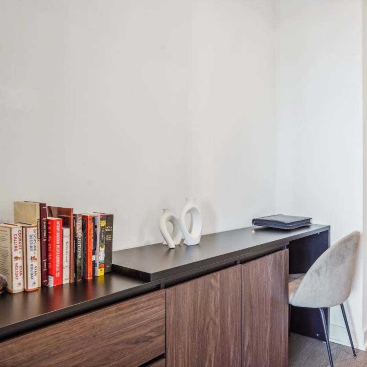 A narrow hallway with a black desk, a chair, a small stack of books, and a white sculpture, leading to a door at the end.