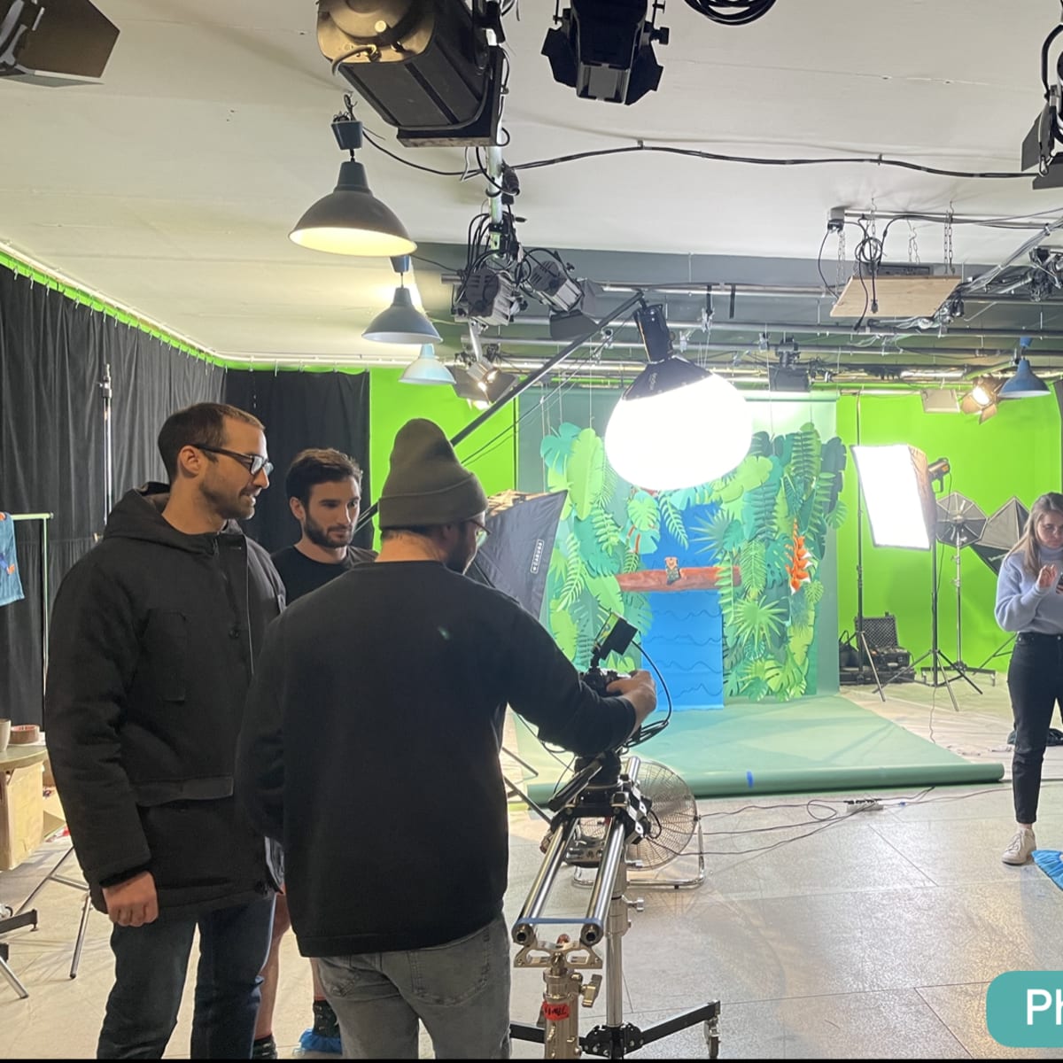 A film crew prepares equipment in a studio with a green screen and various lighting setups around a set designed with tropical foliage.