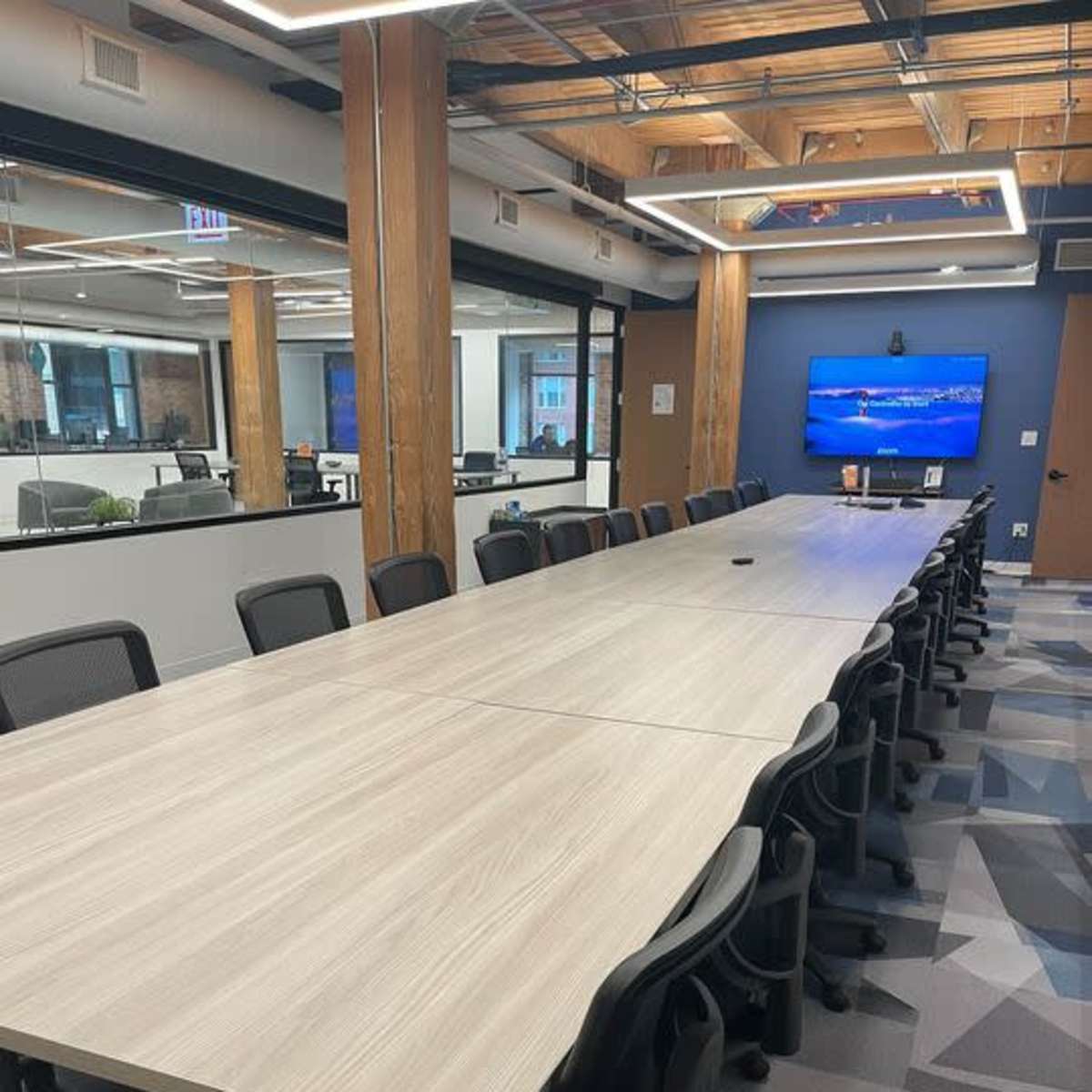 A long wooden conference table is surrounded by black office chairs in a modern office space with a large screen displaying a landscape image.