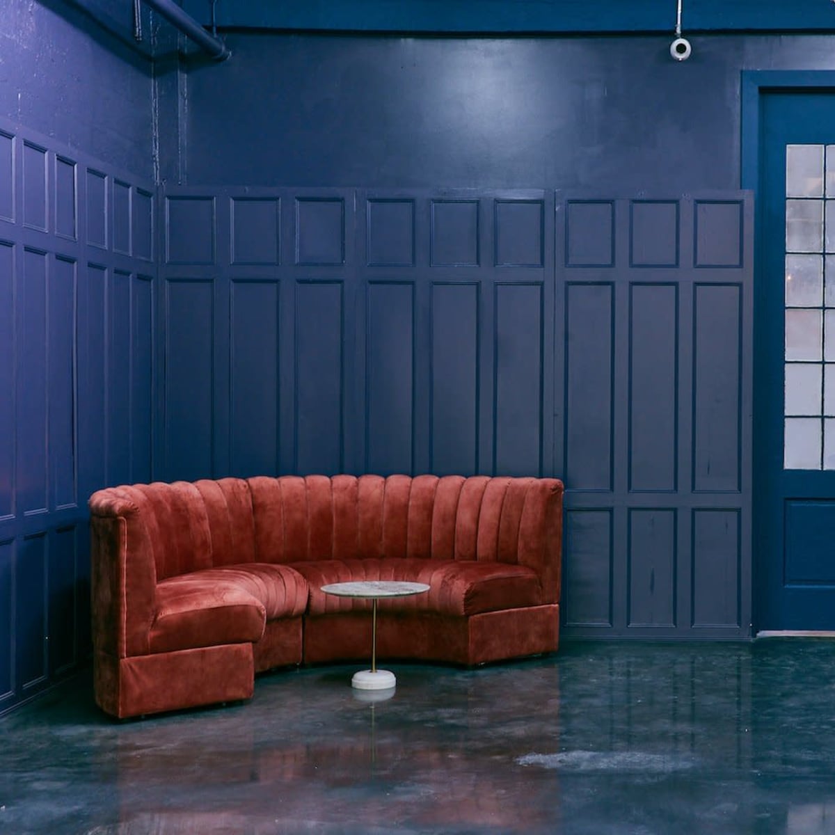 A corner seating area with a curved red velvet sofa and a glass-top coffee table against a dark blue wall featuring large glass double doors.