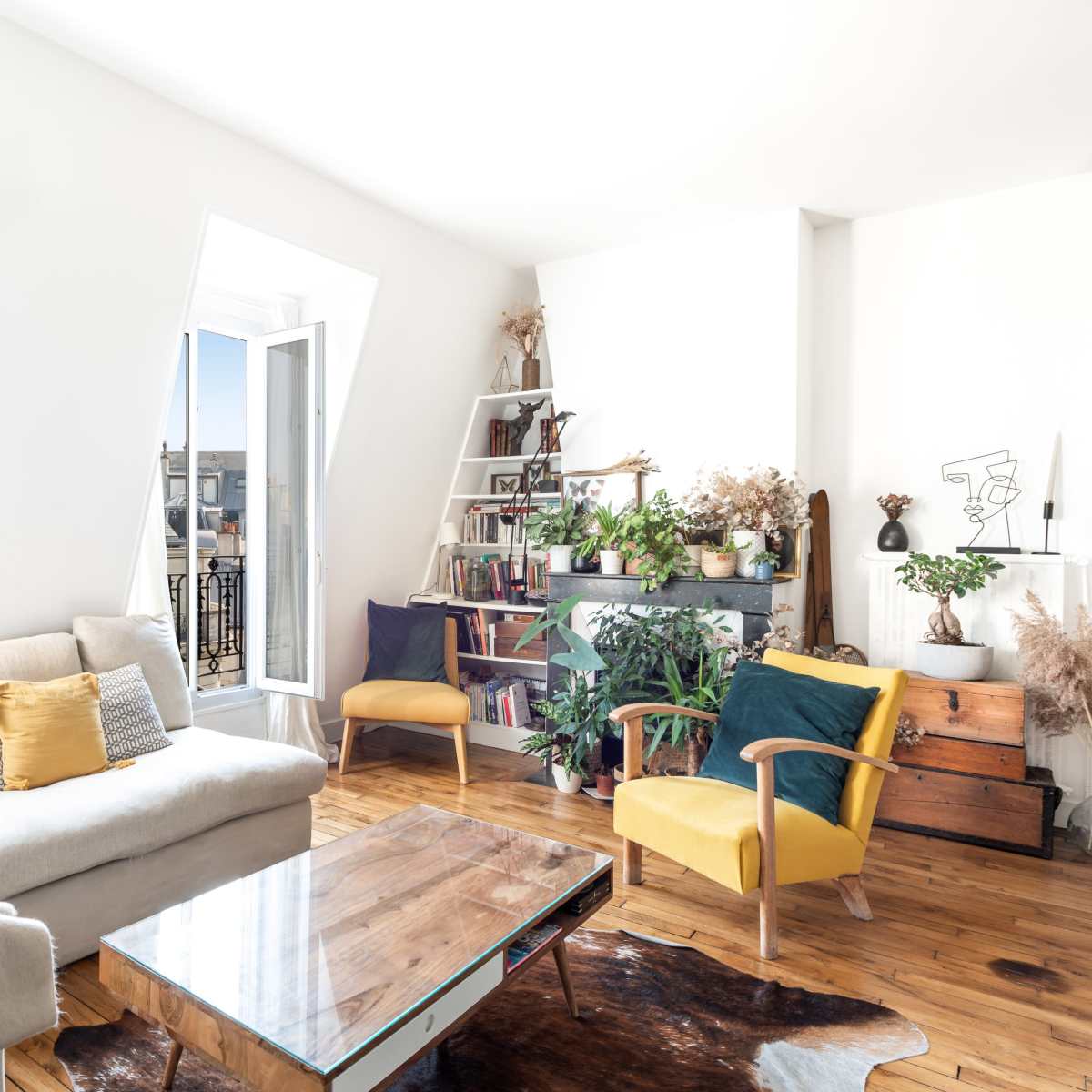 A bright living room featuring a light-colored sofa, a wooden coffee table, and a bookshelf filled with plants and decor, complemented by an open window with a view of rooftops.