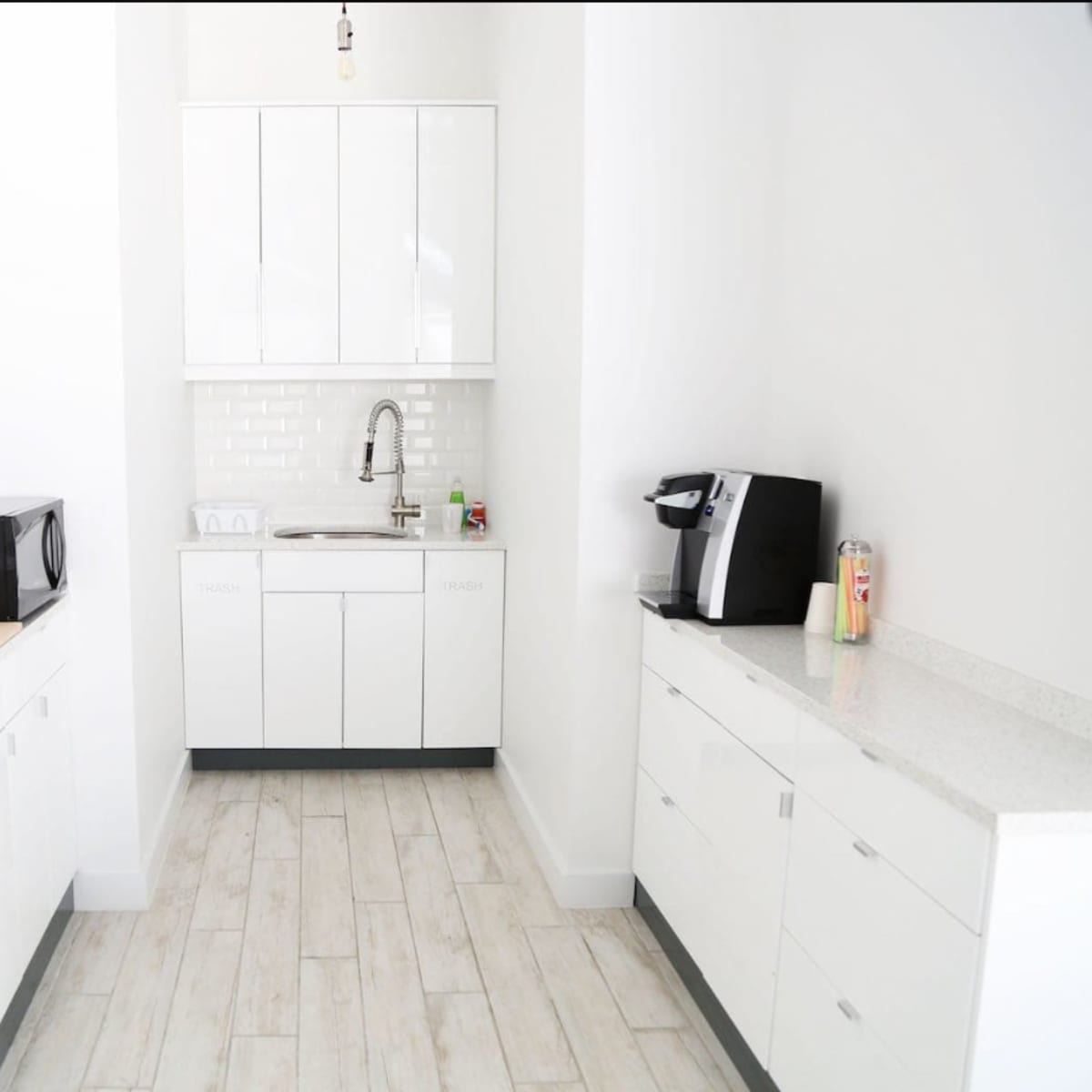 A modern kitchen space featuring white cabinetry, a sink, a microwave, and a coffee machine, with light-colored flooring.