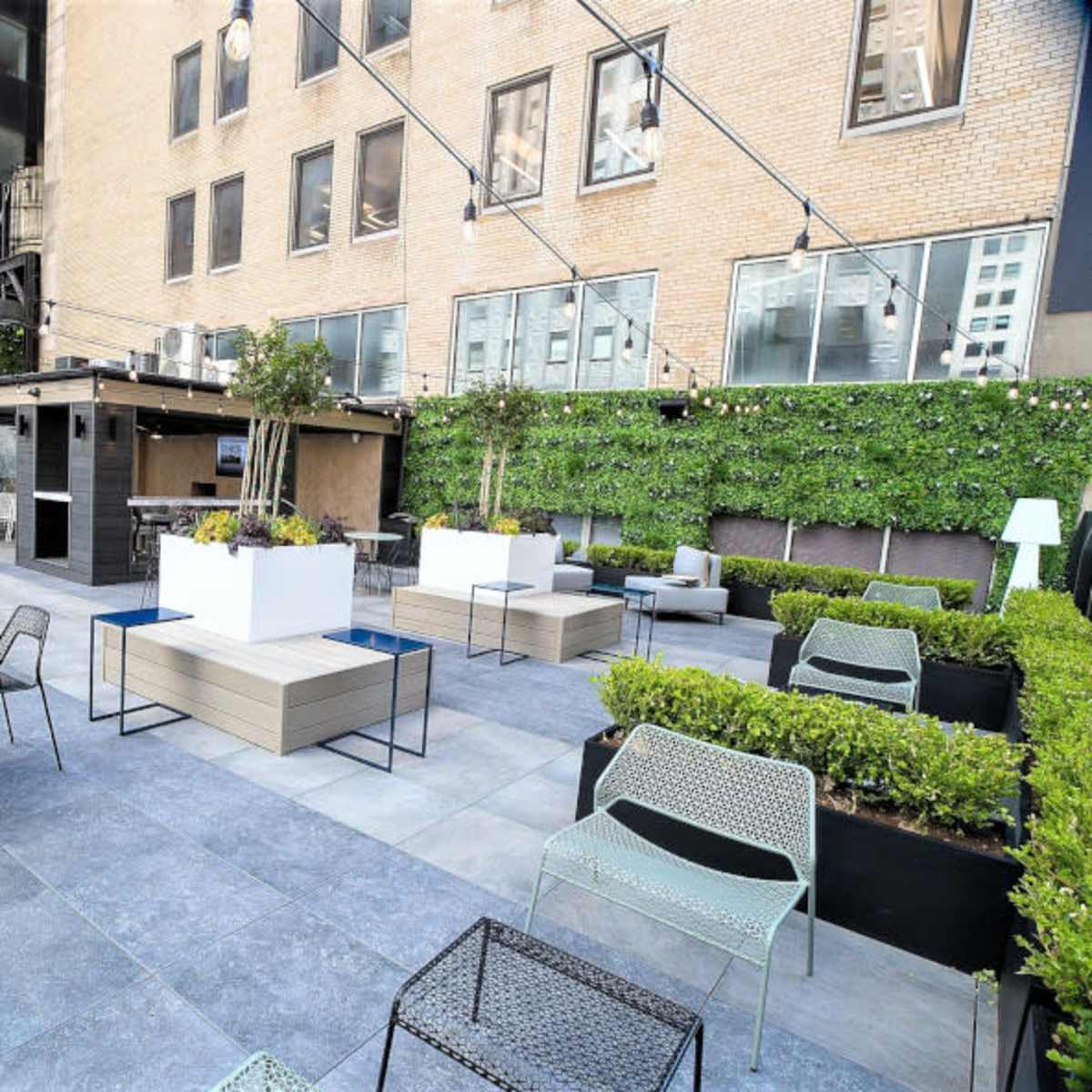 A modern outdoor patio area featuring seating arrangements, planters, and a bar, surrounded by tall buildings.