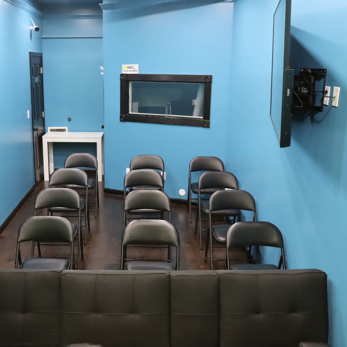 A small room with light blue walls, containing a row of black chairs facing a television and a window.