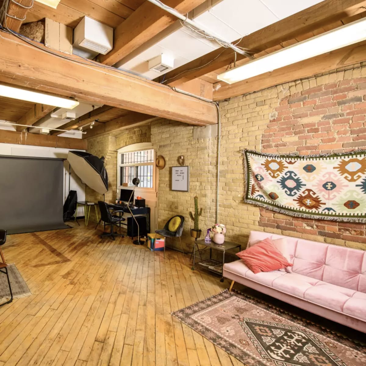 A spacious studio with wooden beams, a large gray backdrop, a pink sofa, and a desk with a chair.