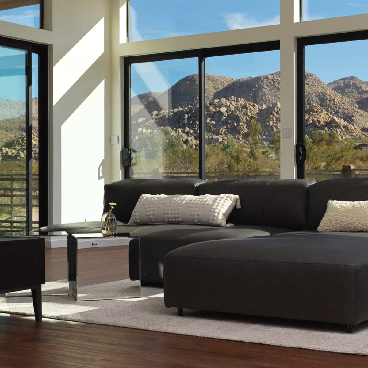 A modern living room features a black sectional sofa and a glass coffee table, with large windows showcasing a mountain landscape.
