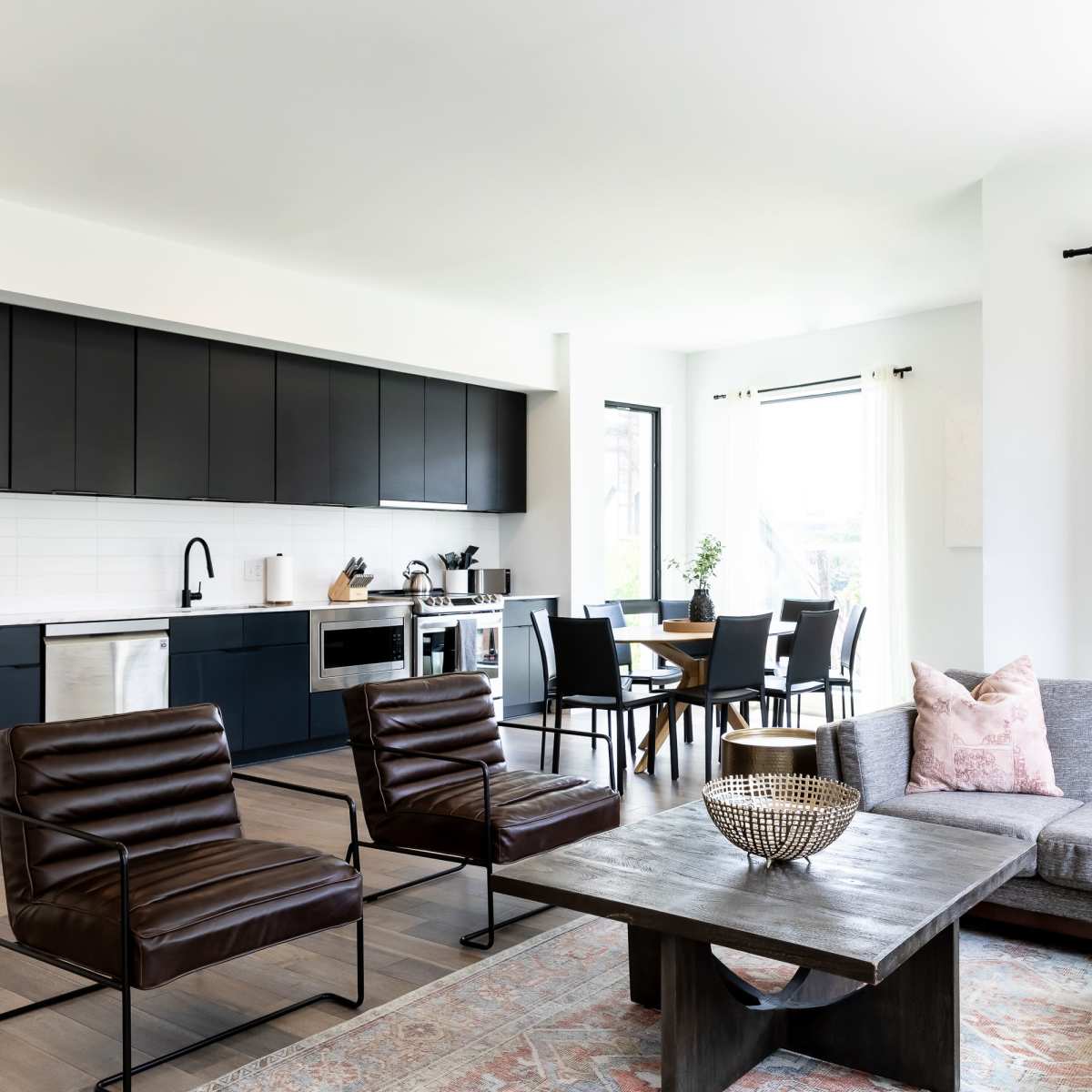 A modern living space features a gray sofa and brown chairs, a wooden coffee table, and a kitchen visible in the background with dark cabinetry.