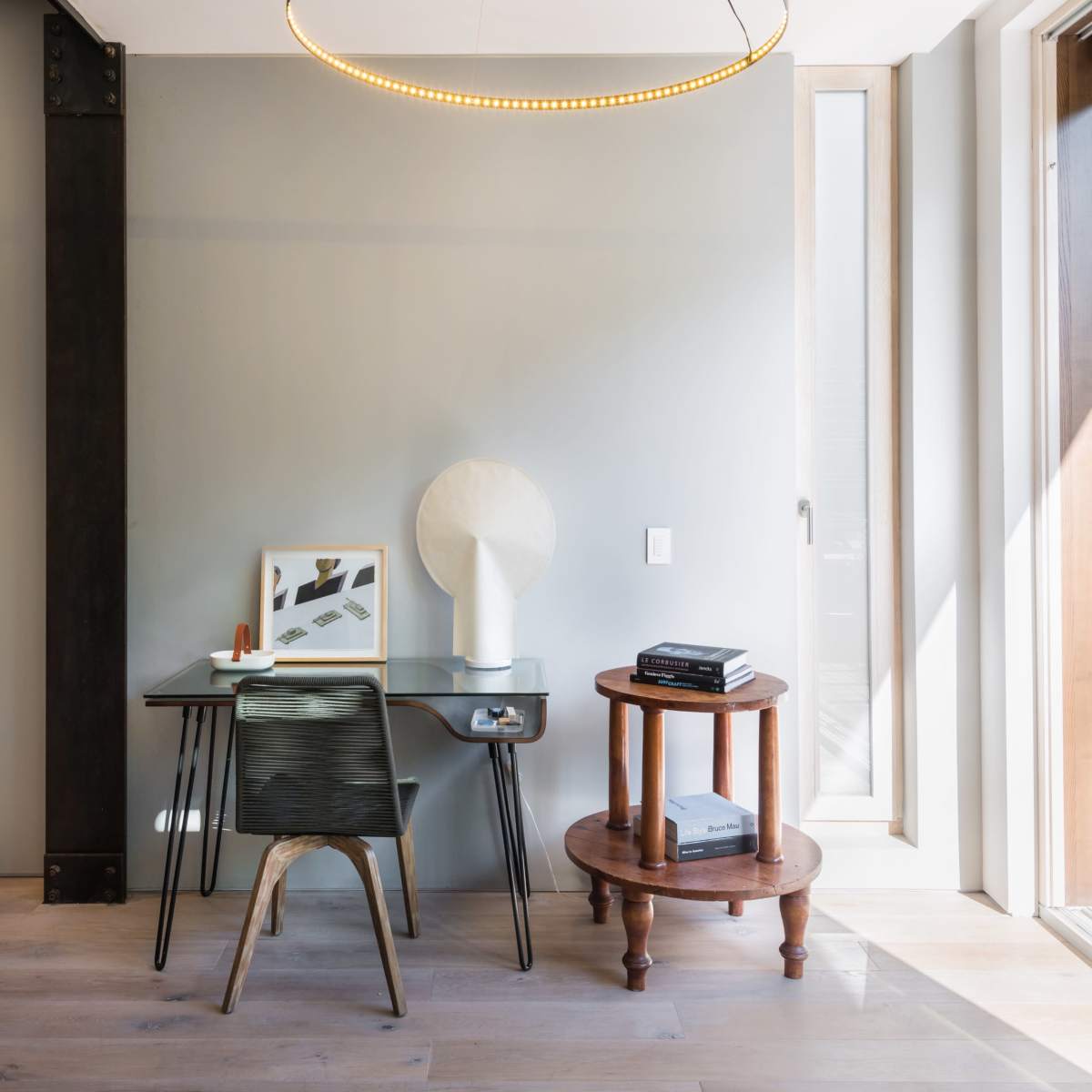 A modern workspace features a round pendant light above a desk with a decorative lamp and artwork, alongside a circular wooden side table.