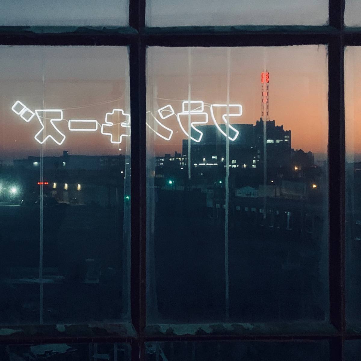 A dimly lit urban scene viewed through a window, featuring glowing text in Japanese characters against a twilight sky.
