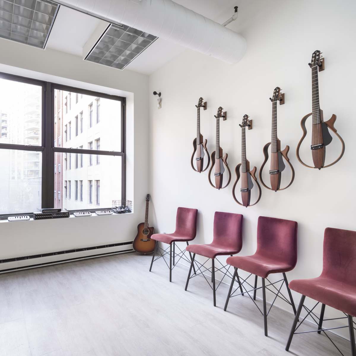 A light-colored room with a window, featuring four maroon chairs in front of an wall adorned with several guitars mounted on hooks.