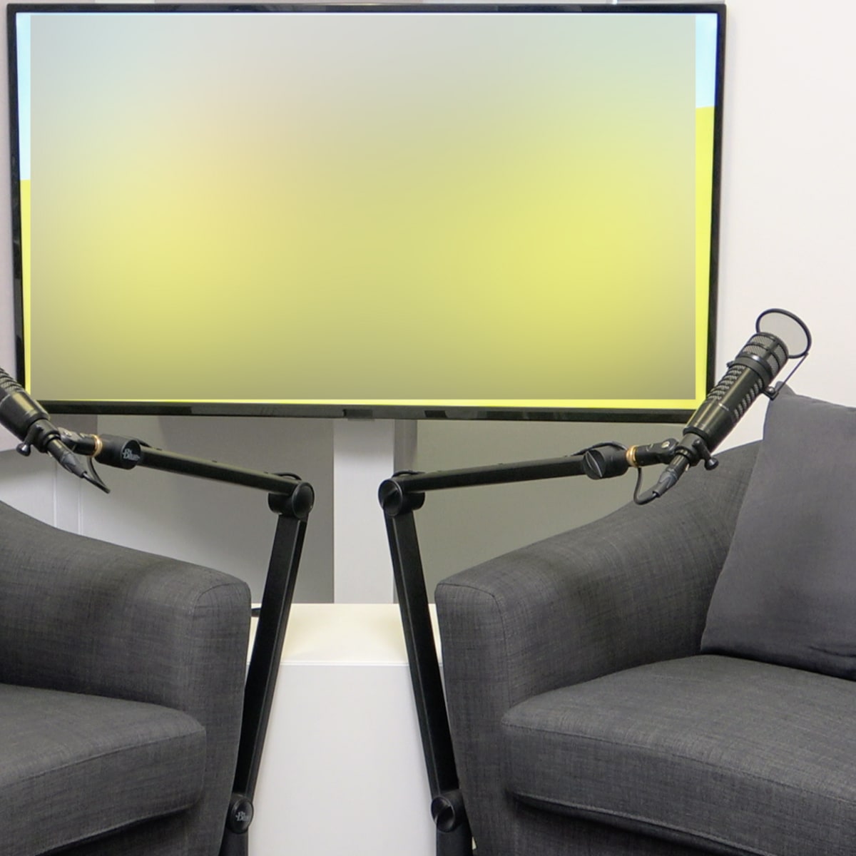 Two gray armchairs positioned in front of a large television screen with a blurred yellow background and two microphones mounted on adjustable arms.