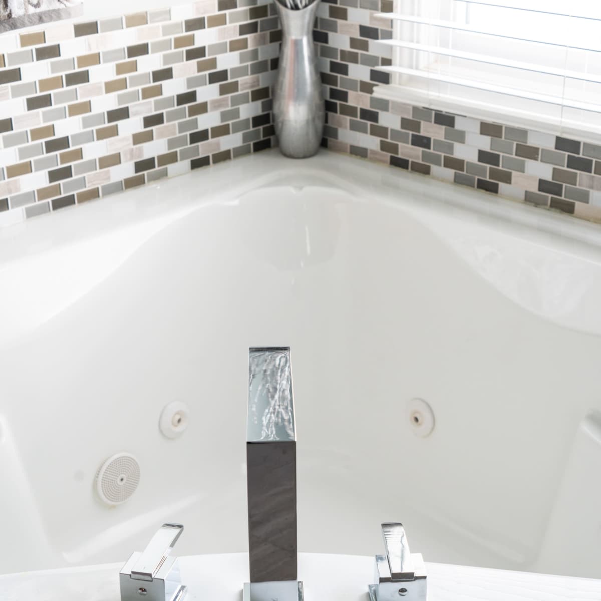 A modern bathtub with three chrome faucets and a mosaic tile backsplash.