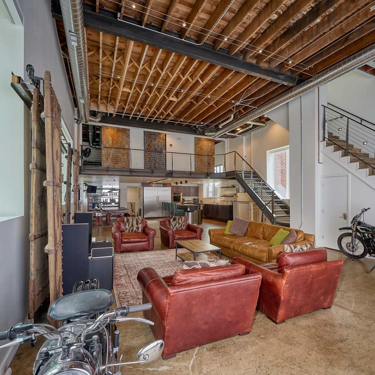 An open-concept living space with exposed wooden beams, multiple seating areas with leather sofas, and a staircase leading to an upper level, along with a motorcycle parked on the floor.