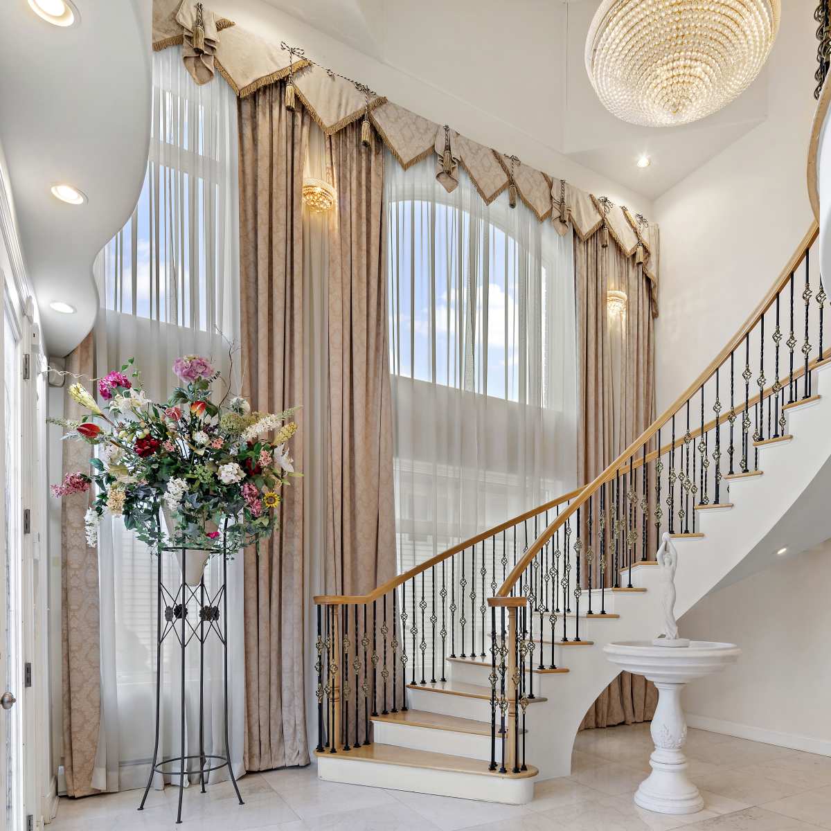 A grand entrance hall features a spiral staircase, large windows, and a floral arrangement on a pedestal.