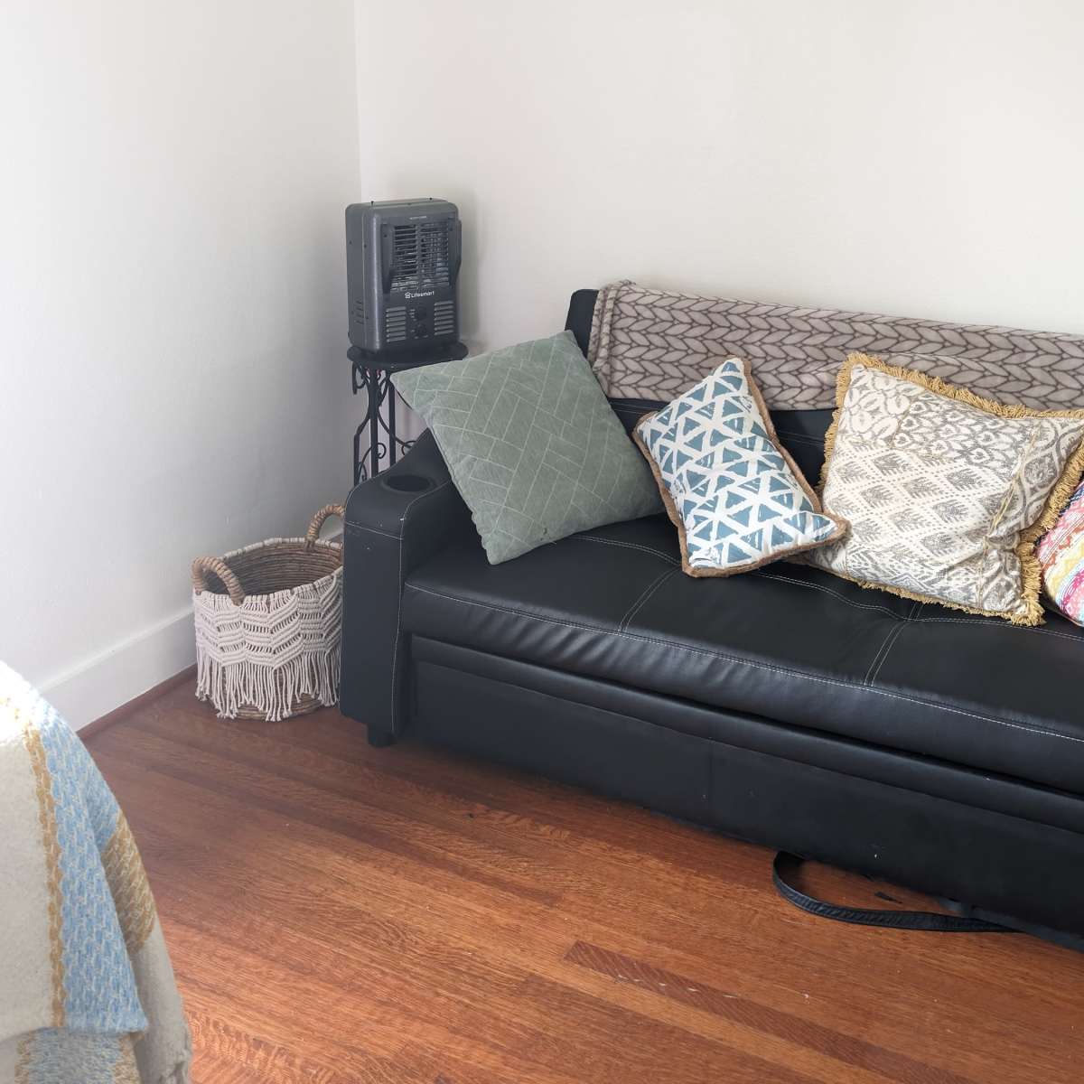 A black sofa with various patterned cushions is positioned against a white wall in a room with wooden flooring.