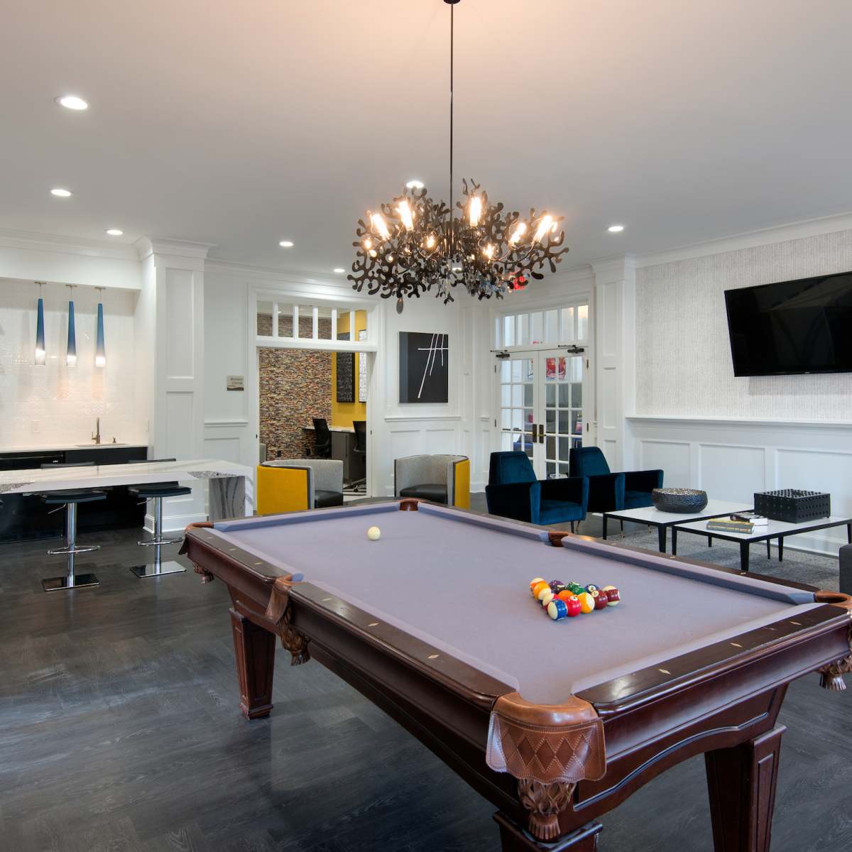 A modern game room featuring a billiard table, a sleek kitchen counter with bar stools, and comfortable seating areas with a chandelier above.