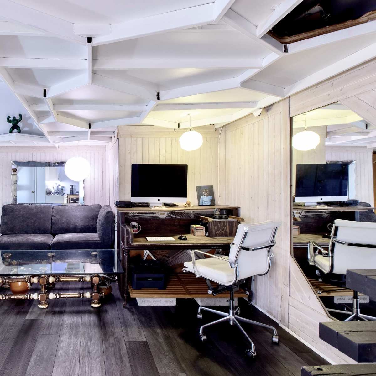 A modern living room with a glass coffee table, a gray sofa, a desk with a computer, and mirrored walls.