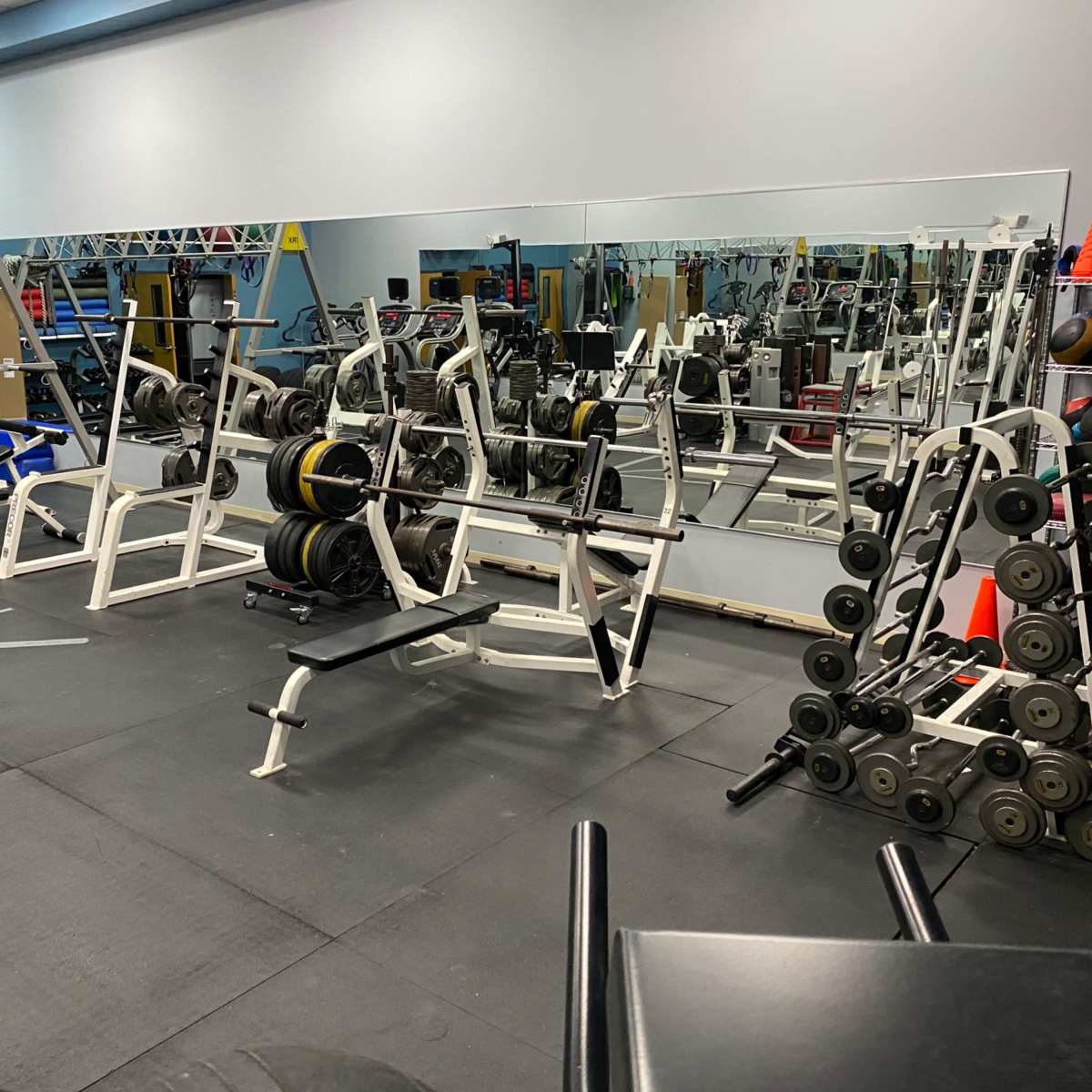 A gym with various weightlifting equipment, including benches and a rack of dumbbells, set against a mirrored wall.