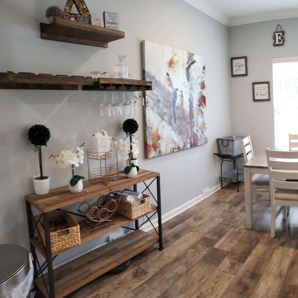 A dining area features a wooden table with white chairs, a shelf with decorative items, and a large window with blinds.