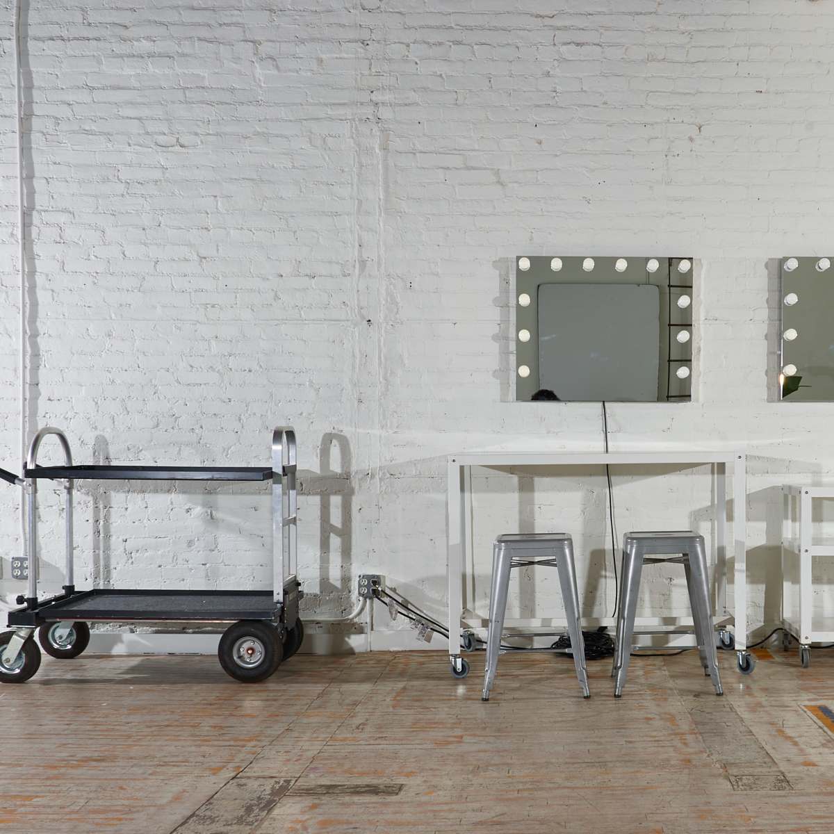 A minimalist interior with a white brick wall, two framed mirrors with lights, a table, two metal stools, a cart, and stacked folding chairs.