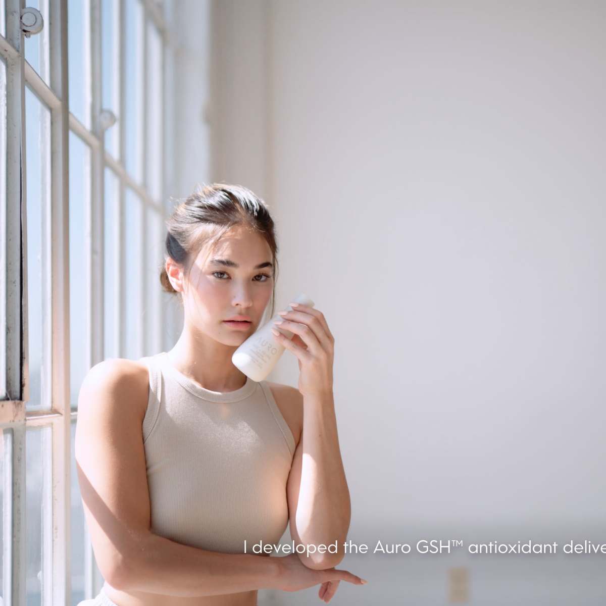 A person stands in a bright, minimalist space while holding a skincare product.