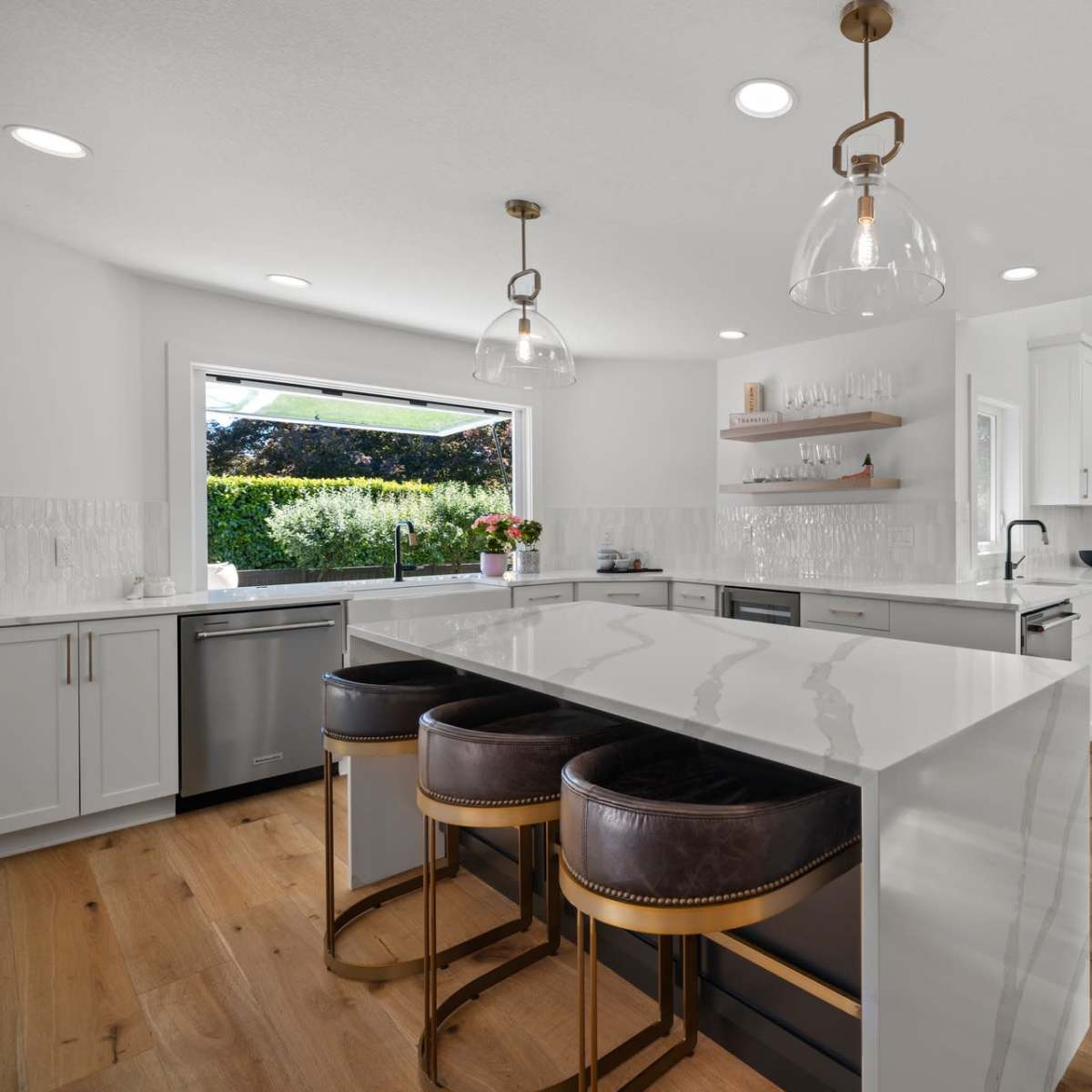 A modern kitchen features a spacious island with three stools, stainless steel appliances, and large windows overlooking a garden.