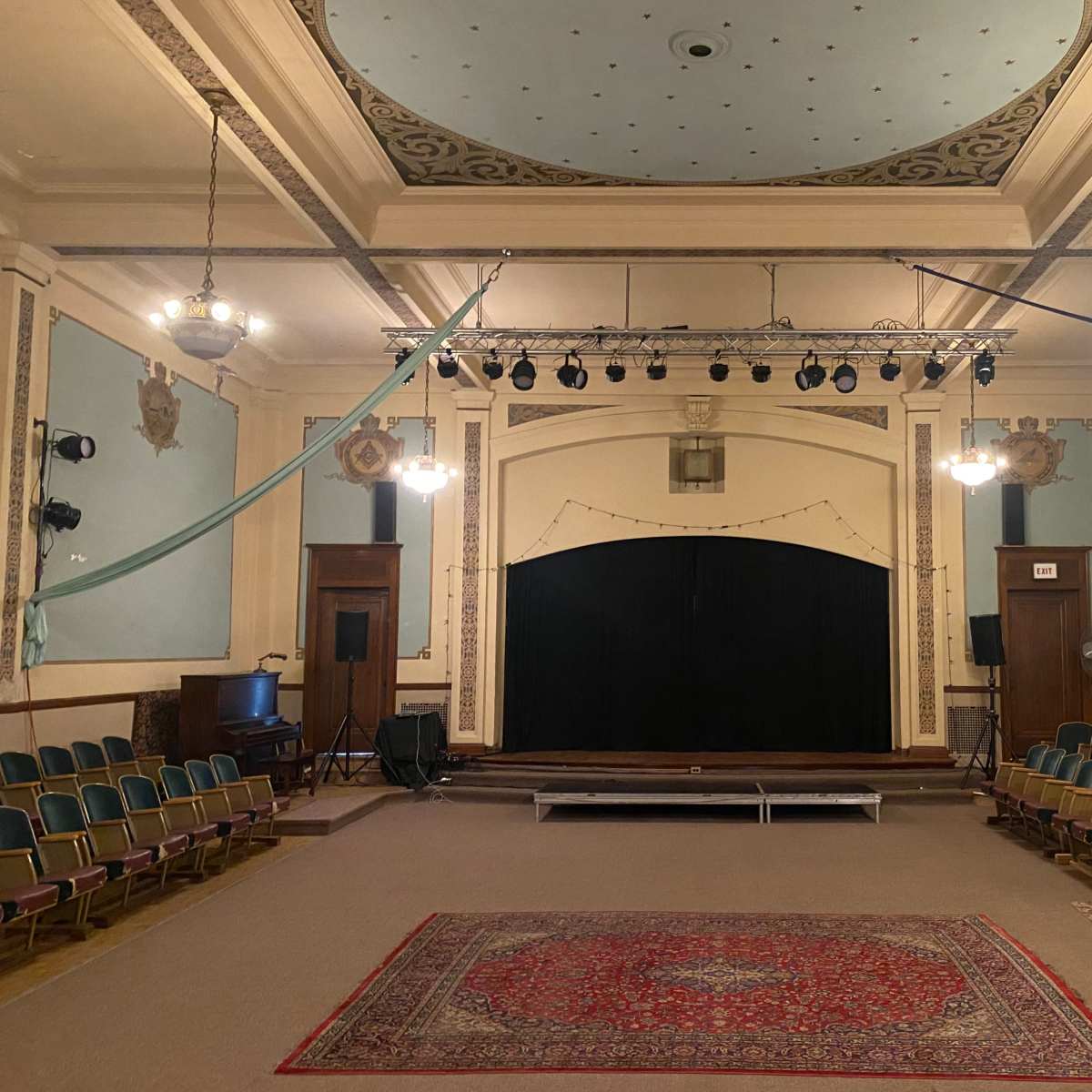 An empty theater with rows of green seats facing a dark stage, illuminated by overhead lights and decorative ceiling fixtures.