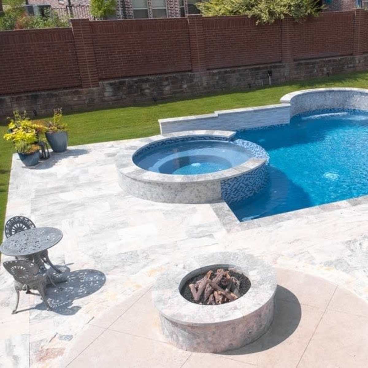 A backyard with a swimming pool, a hot tub, white lounge chairs, and a small fire pit surrounded by stone pavement.