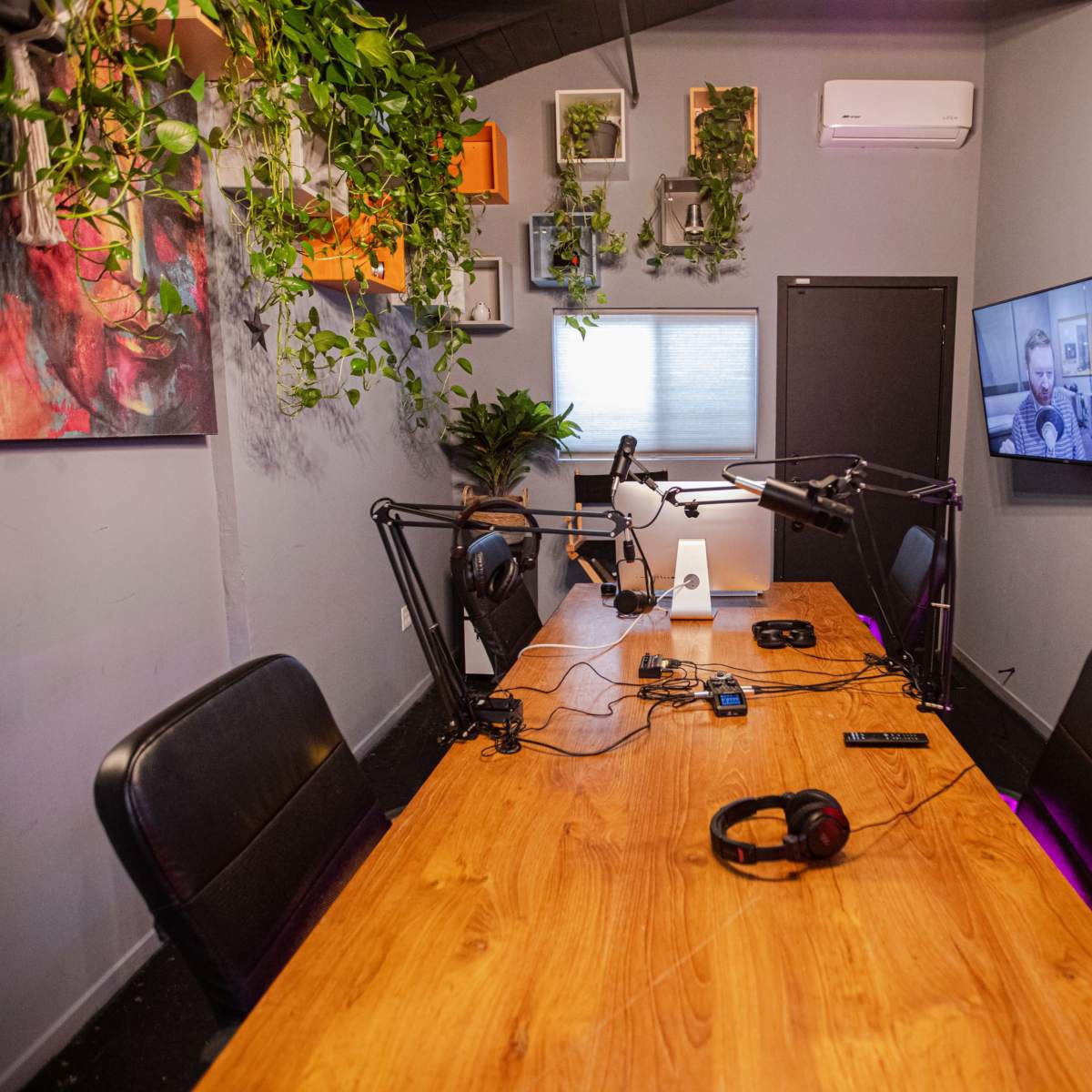 A podcast recording studio features a wooden table with microphone setups, headphones, and wall-mounted greenery.