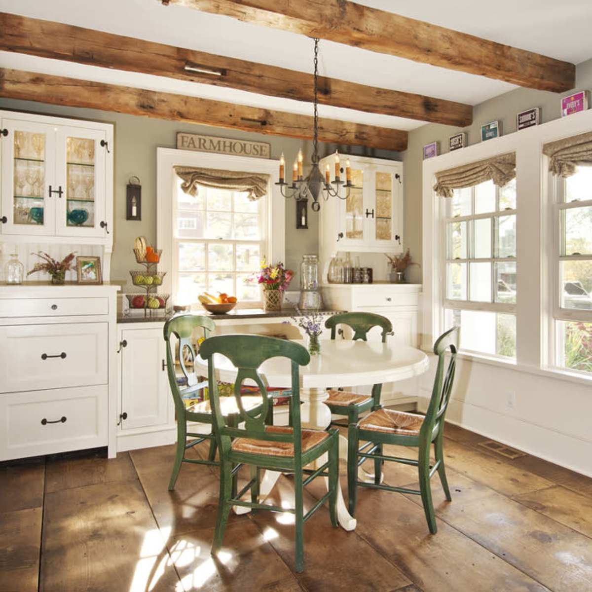 A dining area features a round white table surrounded by green chairs, with wooden beams and large windows illuminating the space.