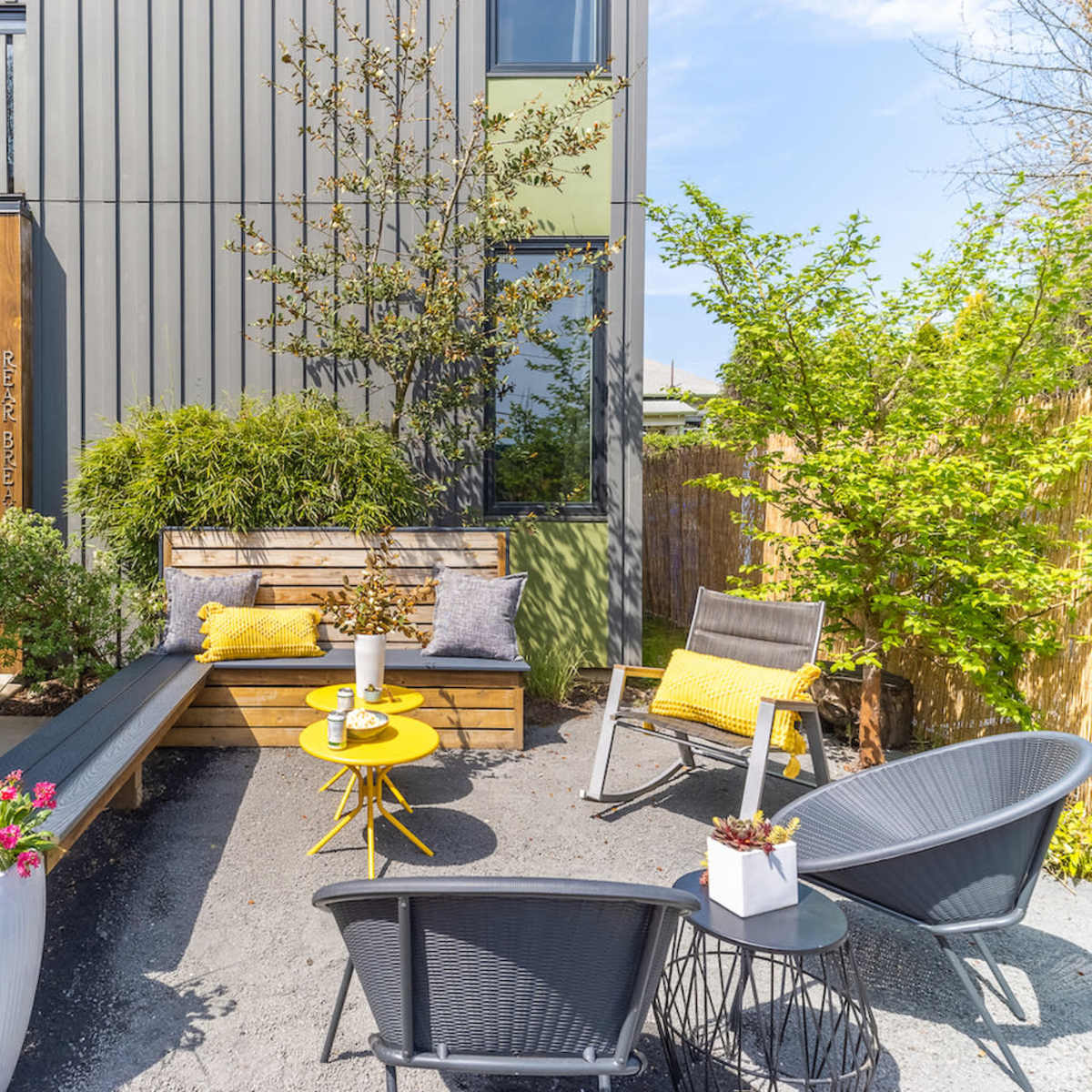 A patio area features modern seating arranged around a yellow table, surrounded by greenery and decorative plants.