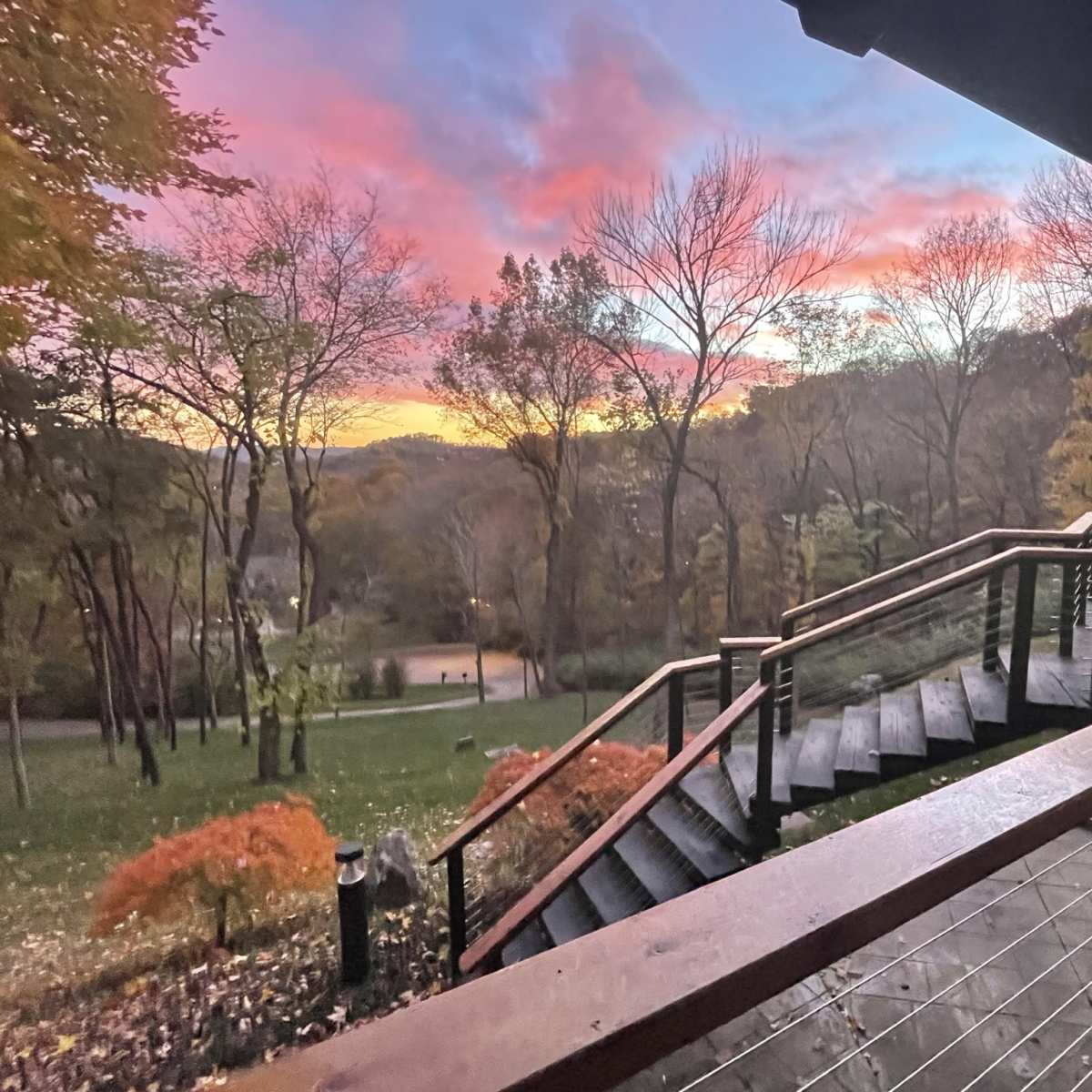 A wooden staircase leads down from a balcony, offering a view of a sunset over a valley filled with trees.