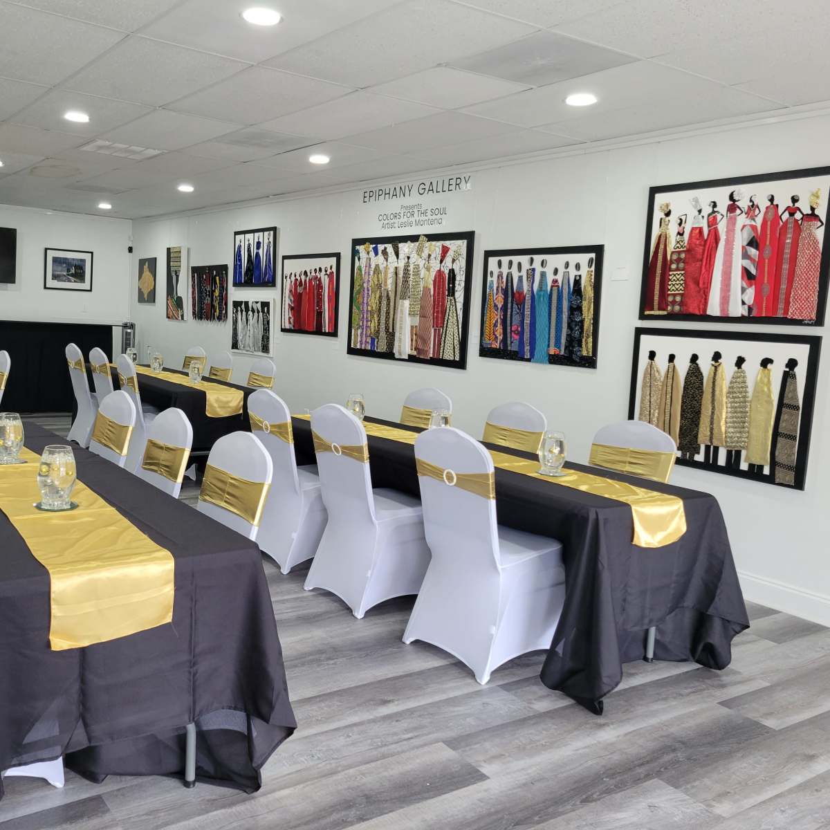 A room set up for an event with several long tables covered in black and gold tablecloths, surrounded by white chairs, and adorned with framed garments on the walls.