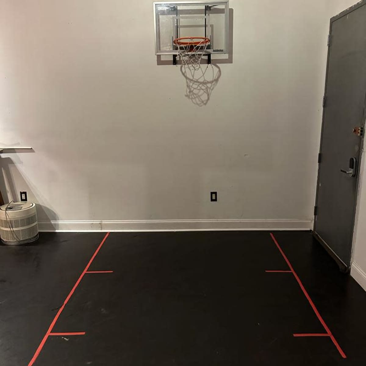 A basketball hoop is mounted on the wall above a marked court area with red tape on the floor.