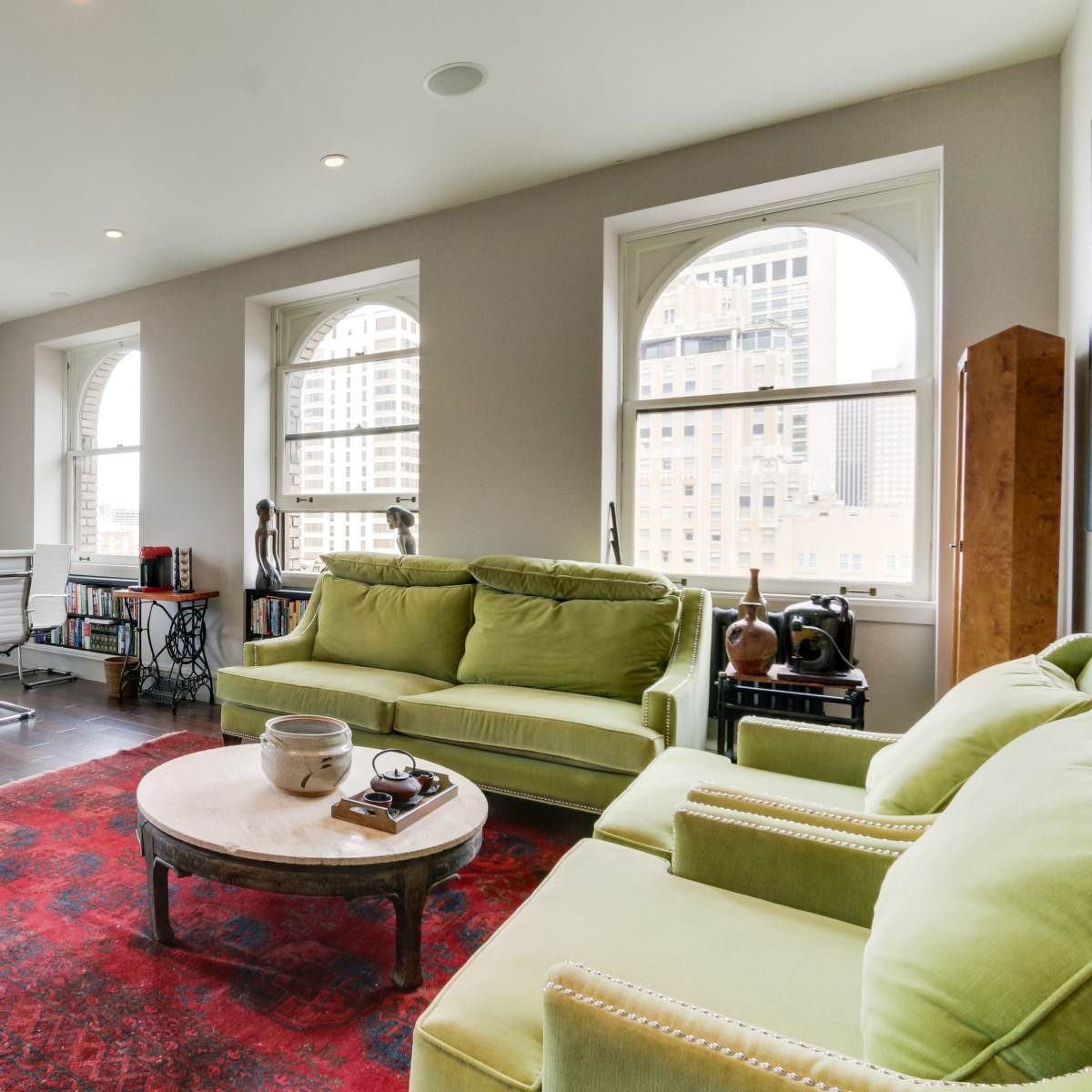 A modern living space features a green sofa, a round coffee table, and a workspace with white chairs, all set against large arched windows.