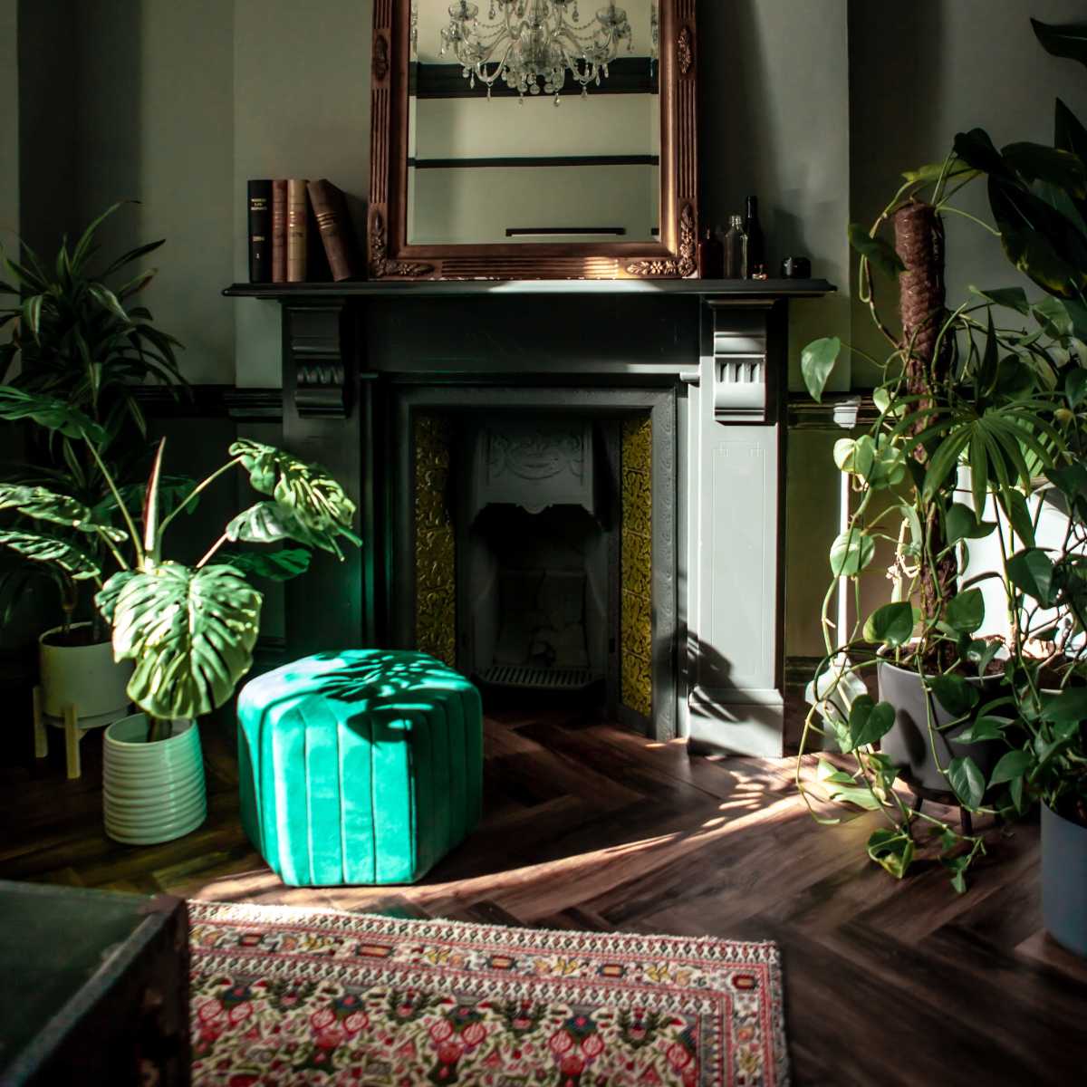 A room with a dark fireplace, a large mirror above it, green plants around, and a green upholstered ottoman on a patterned rug.
