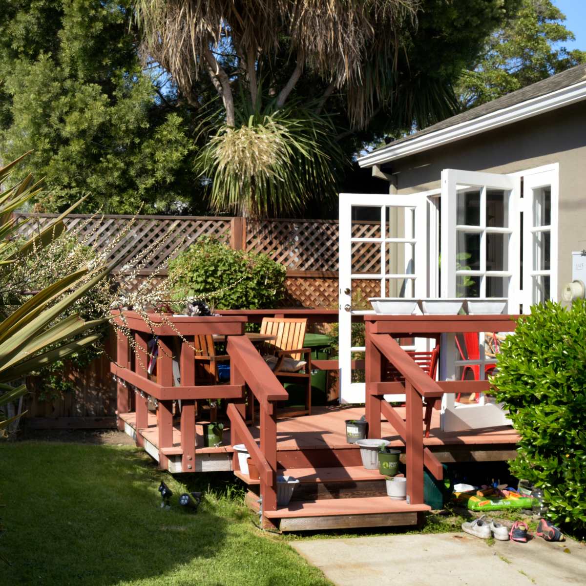 A wooden deck with chairs and a table extends from a house, surrounded by greenery and plants in a backyard.