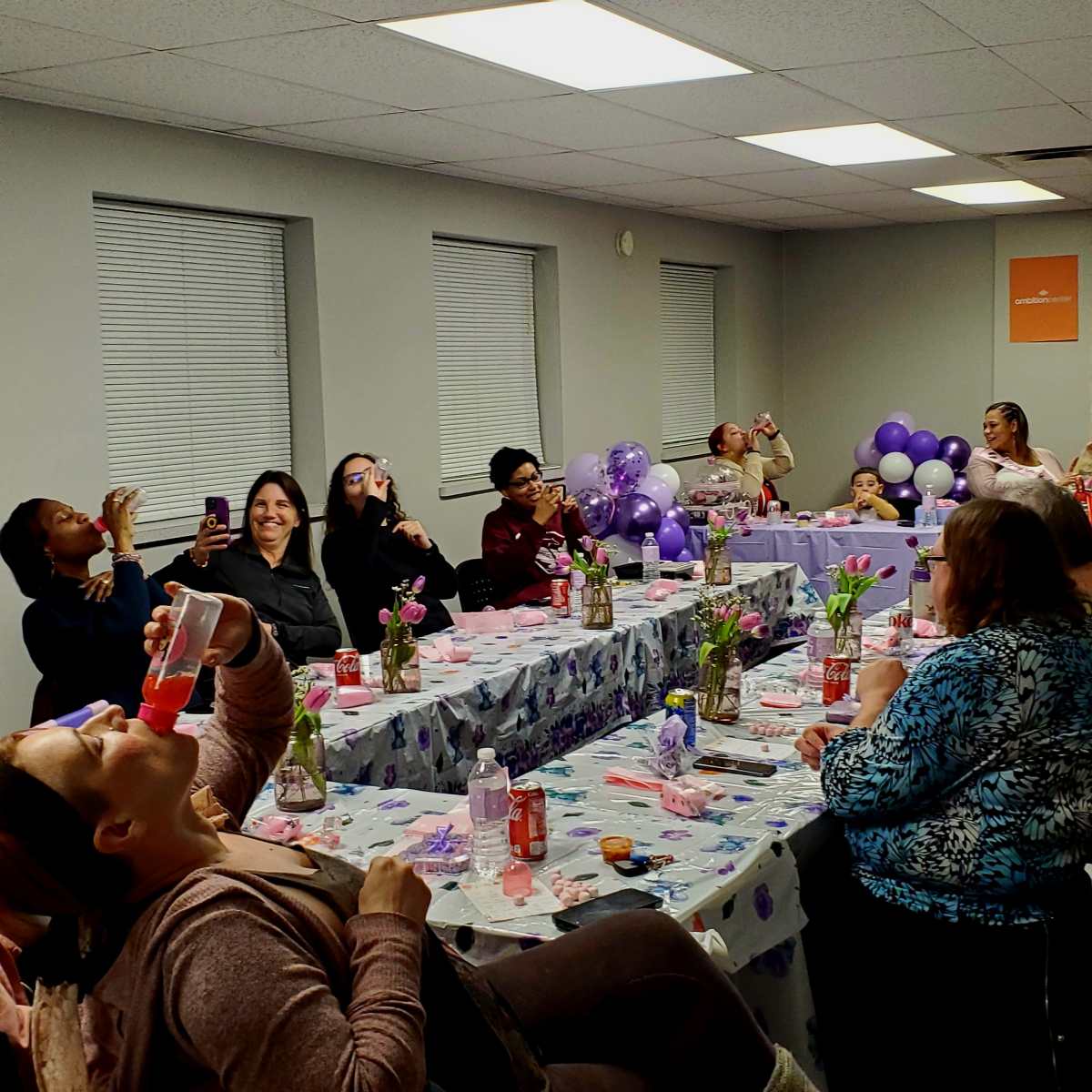 A group of people is sitting at a long table decorated with flowers and balloons, enjoying drinks and engaging in conversation.