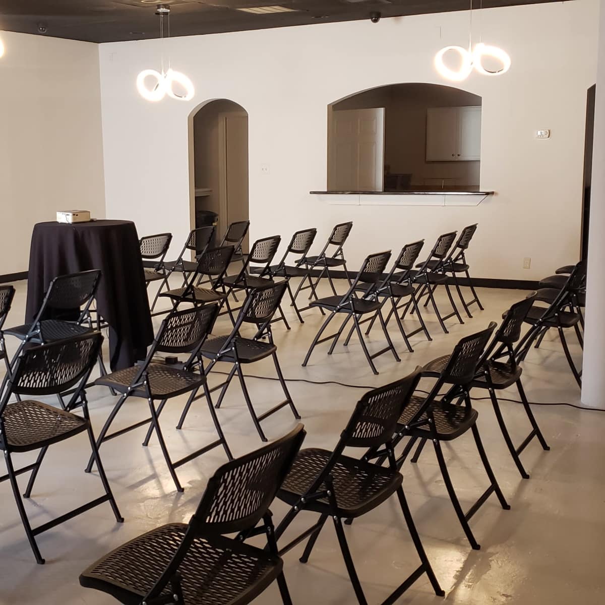 A conference room setup with rows of black folding chairs arranged around a small table and a bar area in the background.