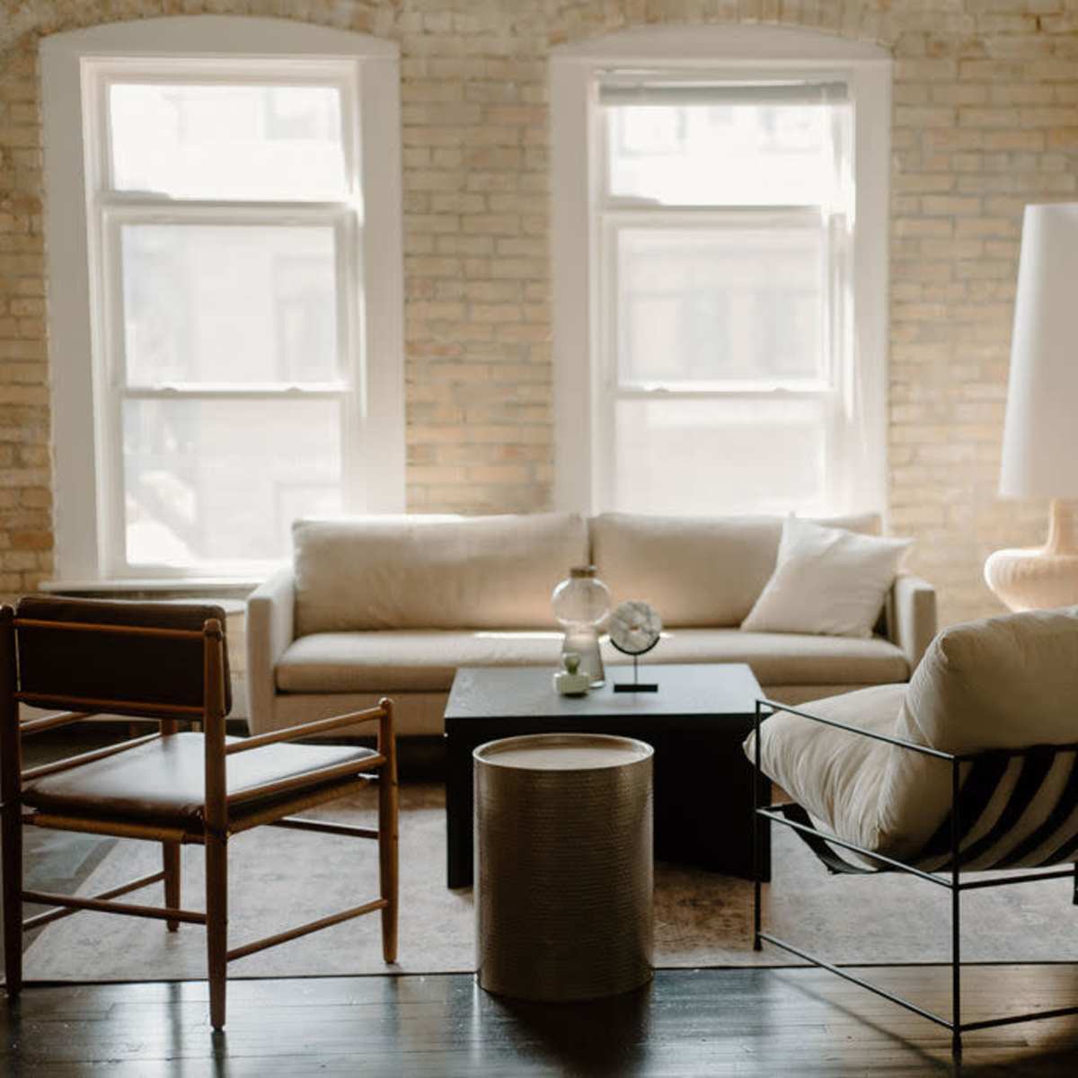 A stylish living room with a beige sofa, a black coffee table, and various decorative elements against a backdrop of exposed brick walls and large windows.