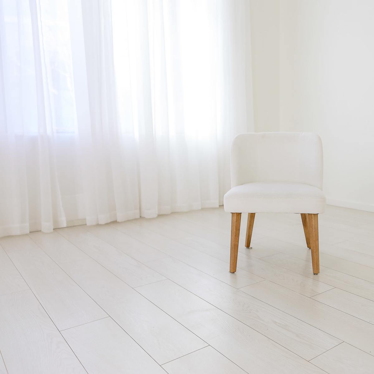 A single white chair stands on a light-colored wooden floor in a bright, minimalist room with sheer white curtains.