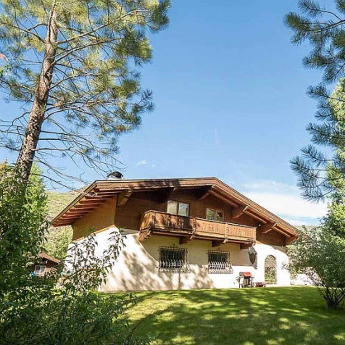 A large house with a wooden balcony sits amid green trees and grass under a clear blue sky.